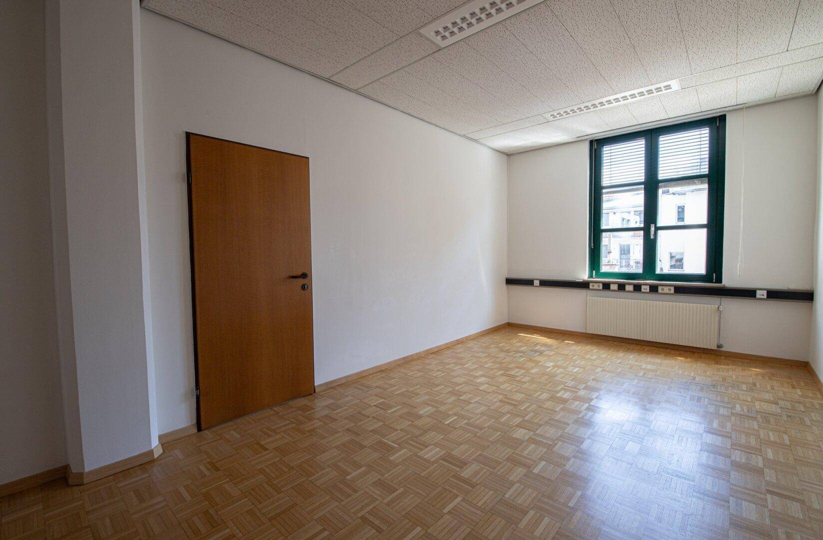 Leeres Büro mit Holztür, Parkettboden und einem Fenster mit Jalousien, das Tageslicht hereinlässt.