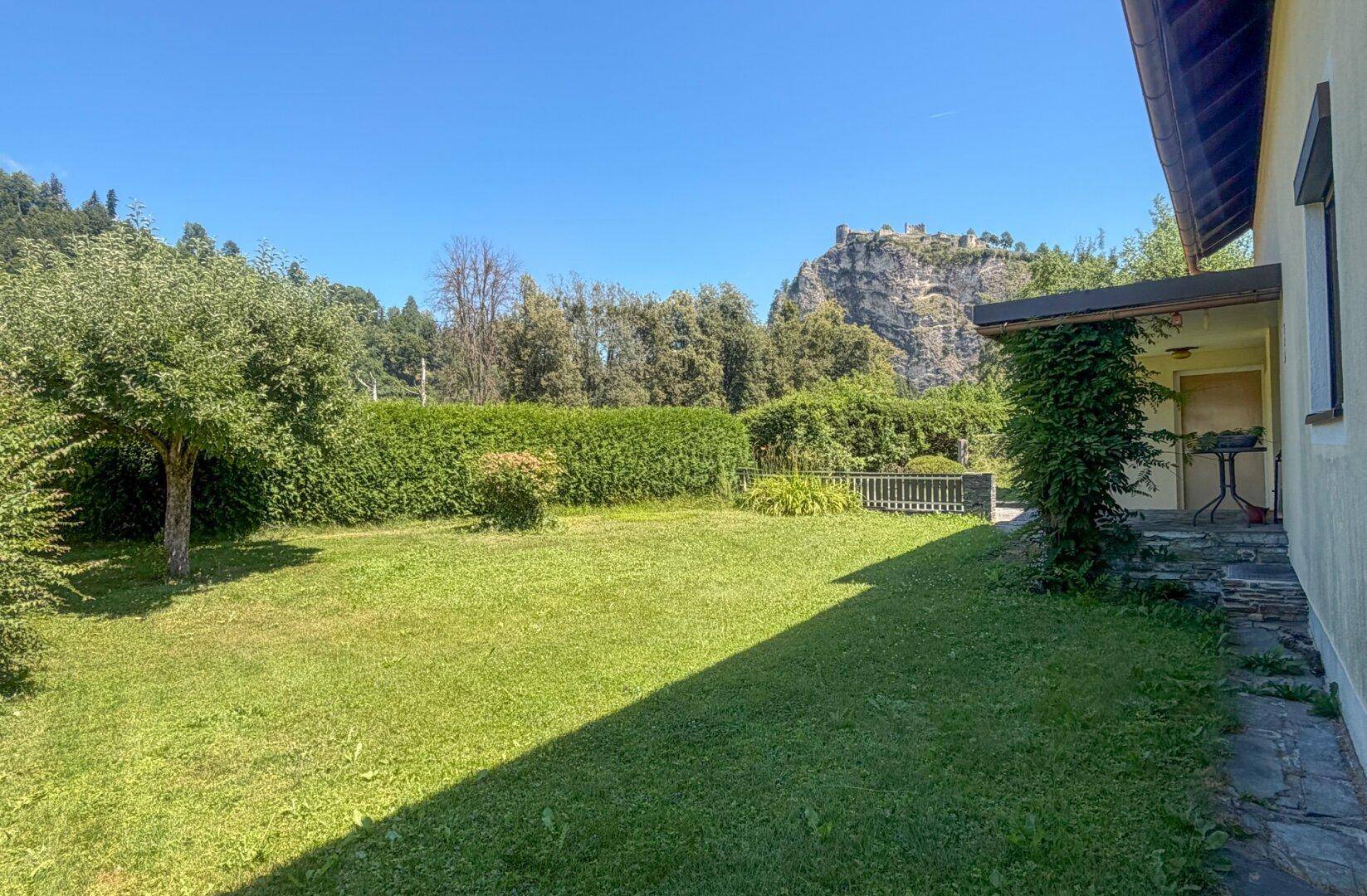 Seitlicher Gartenbereich mit hoher Hecke und Blick auf die umliegende Natur.