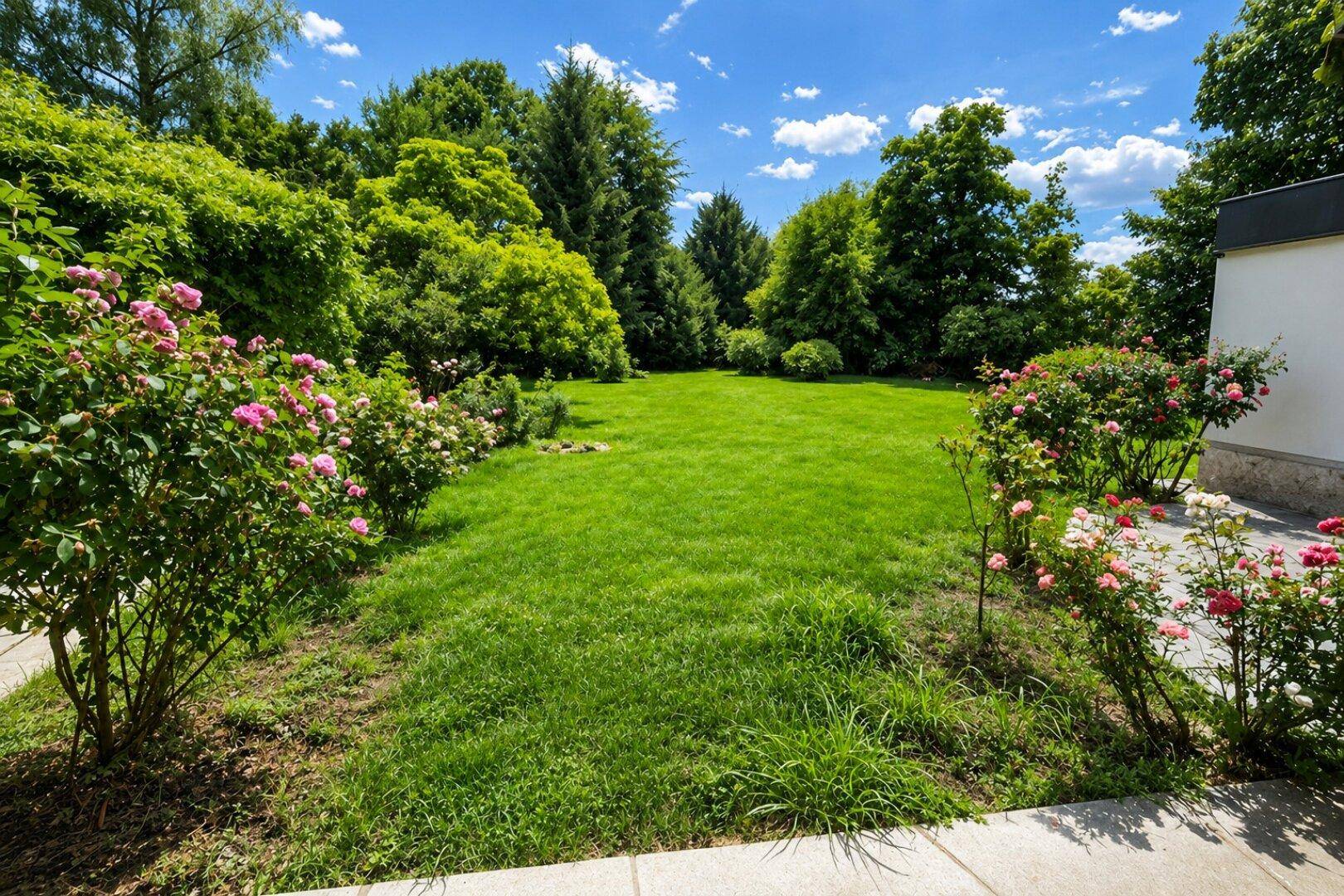 Weitläufiger, gepflegter Garten mit großer Rasenfläche, blühenden Rosenbeeten und altem Baumbestand.