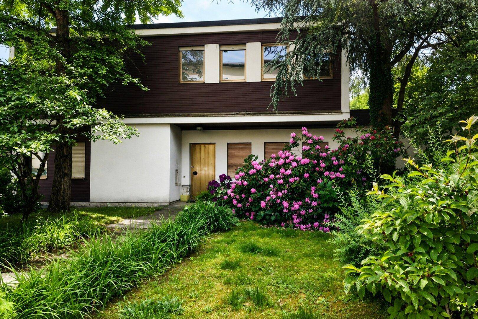 Vorderansicht des Hauses mit gepflegtem Vorgarten, blühenden Rhododendren und altem Baumbestand.