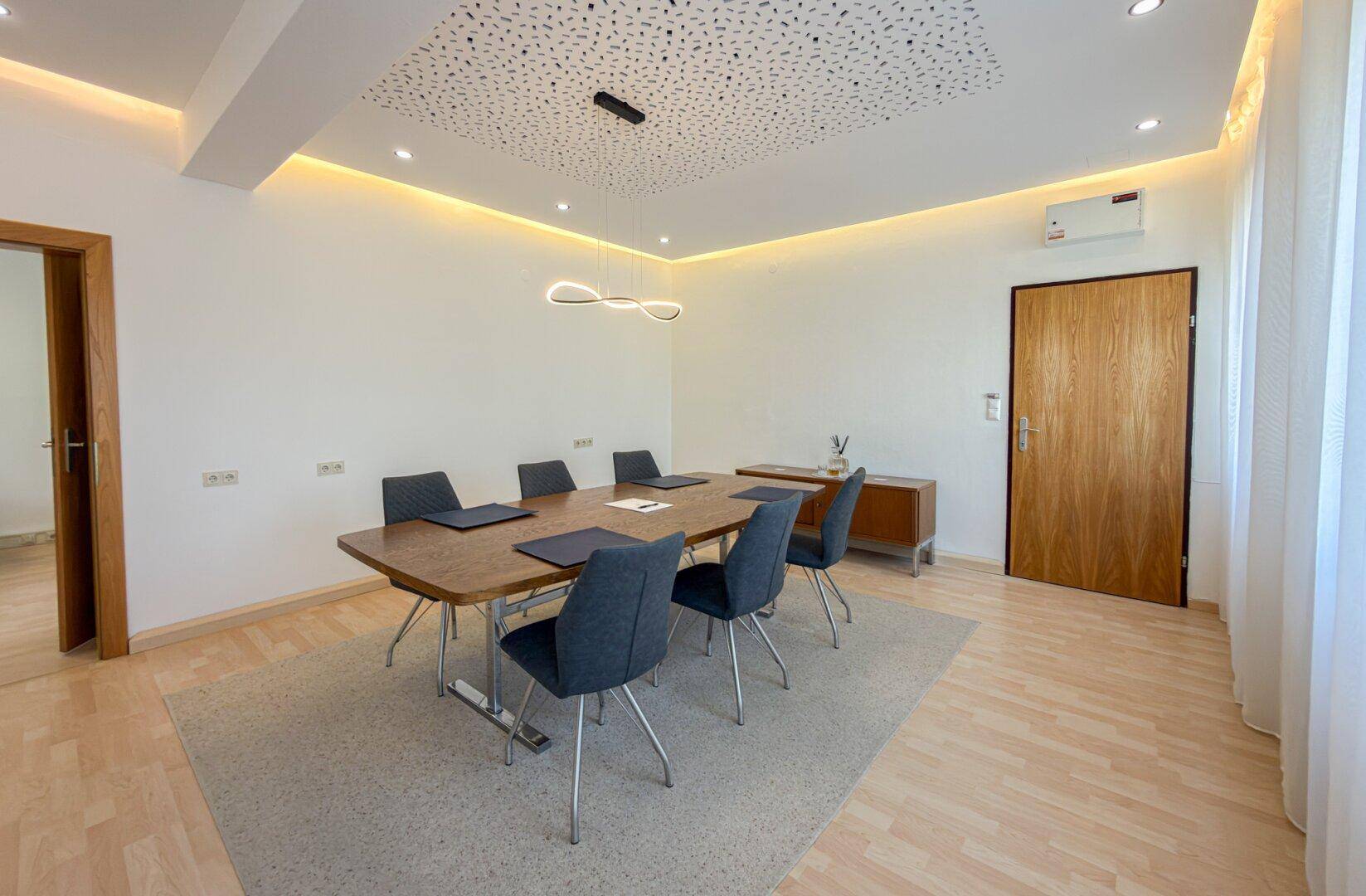 Eleganter Konferenzraum mit Holztisch, grauen Stühlen und einer modernen Deckenbeleuchtung.