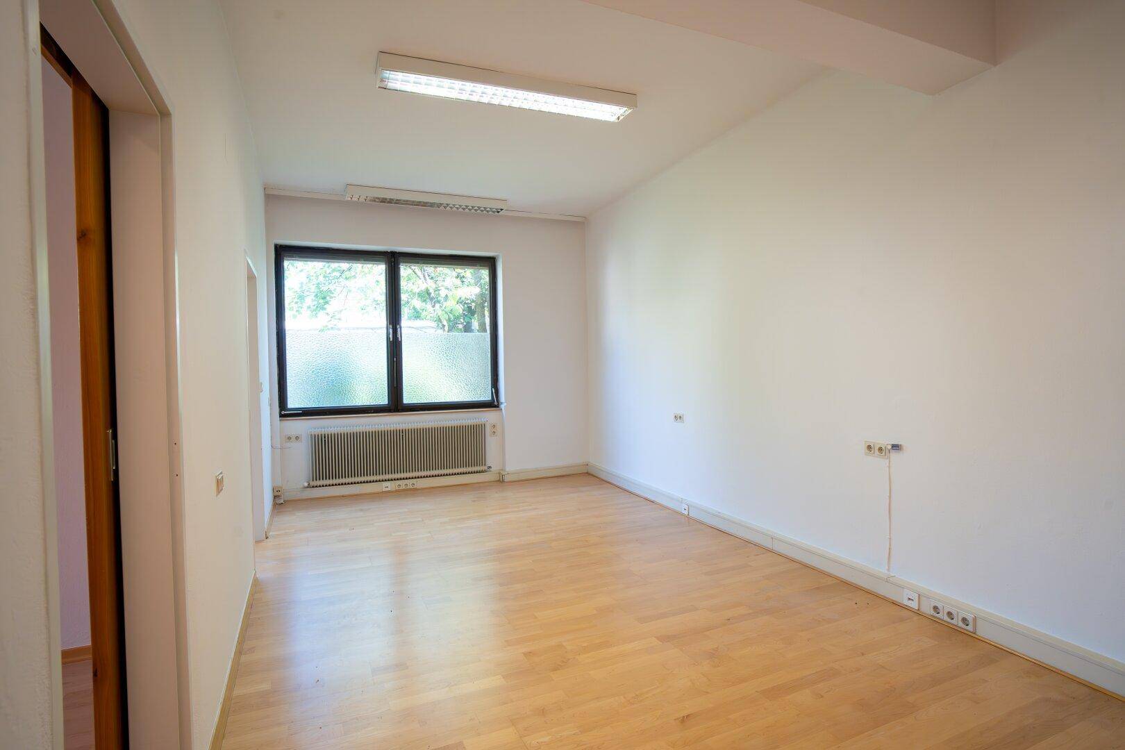 Leeres Büro mit hellem Laminatboden, einem Fenster mit Milchglasfolie und Heizkörper unter dem Fenster.
