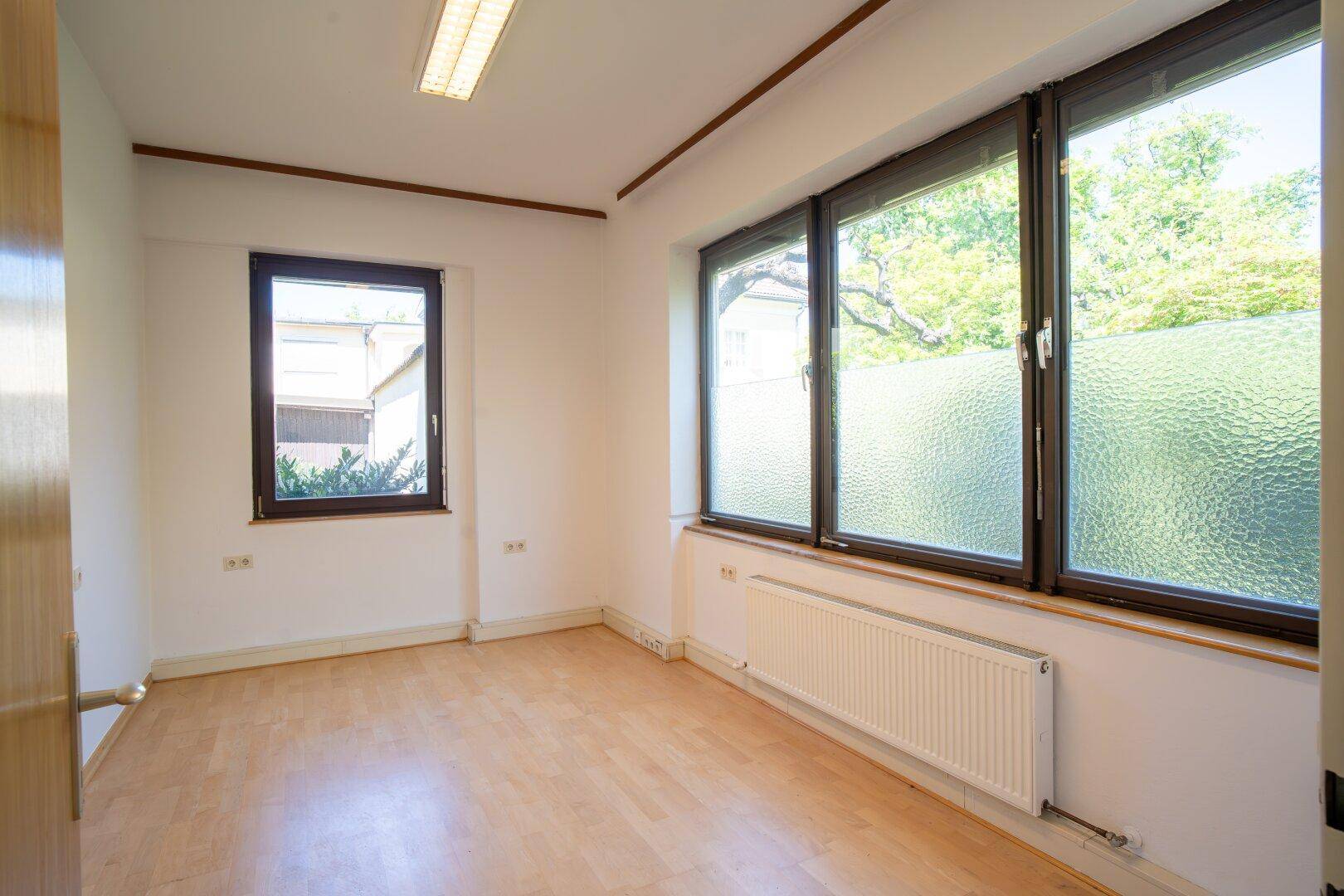 Leeres Büro mit hellem Laminatboden, mehreren Fenstern und einem Heizkörper darunter.
