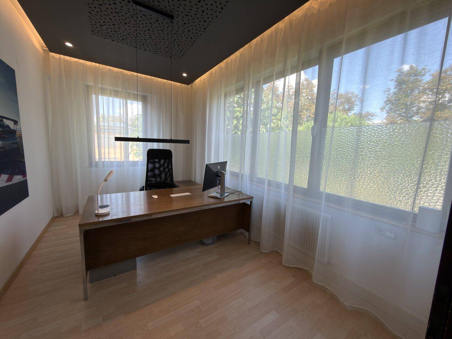 Modernes Büro mit großem Schreibtisch, hellen Vorhängen und einer einzigartigen Deckenbeleuchtung.