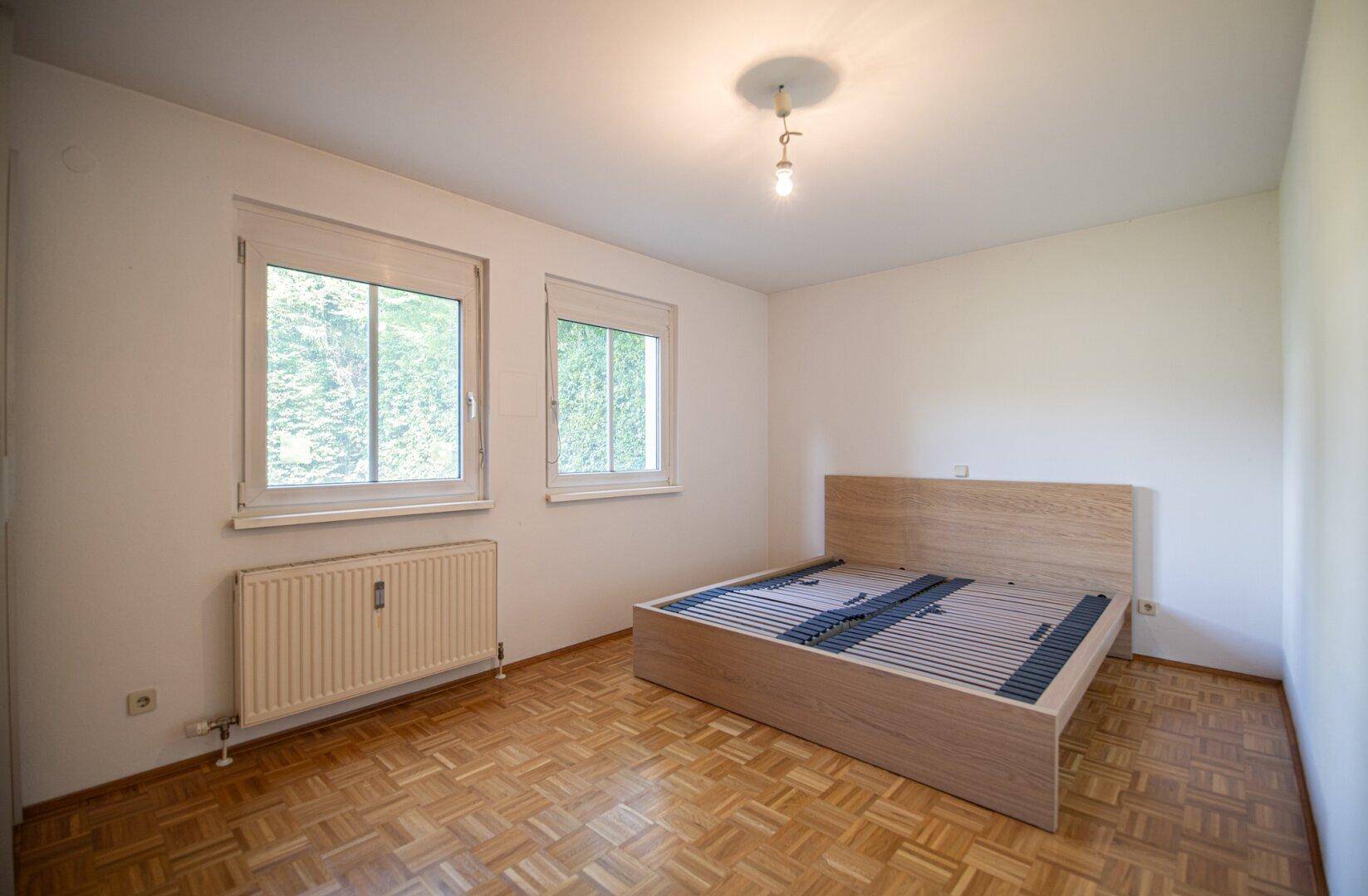 Helles Schlafzimmer mit Parkettboden, einem Holzbettrahmen und zwei Fenstern mit Blick ins Grüne.