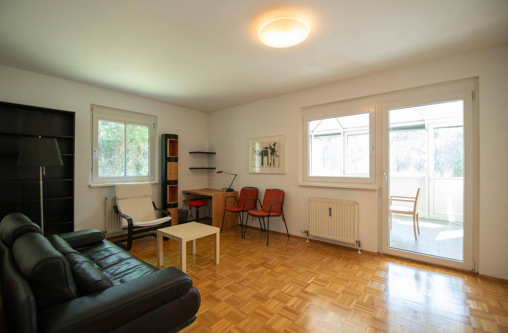 Wohnzimmer mit Parkettboden, schwarzem Sofa und Zugang zu einem sonnigen Balkon oder Wintergarten.