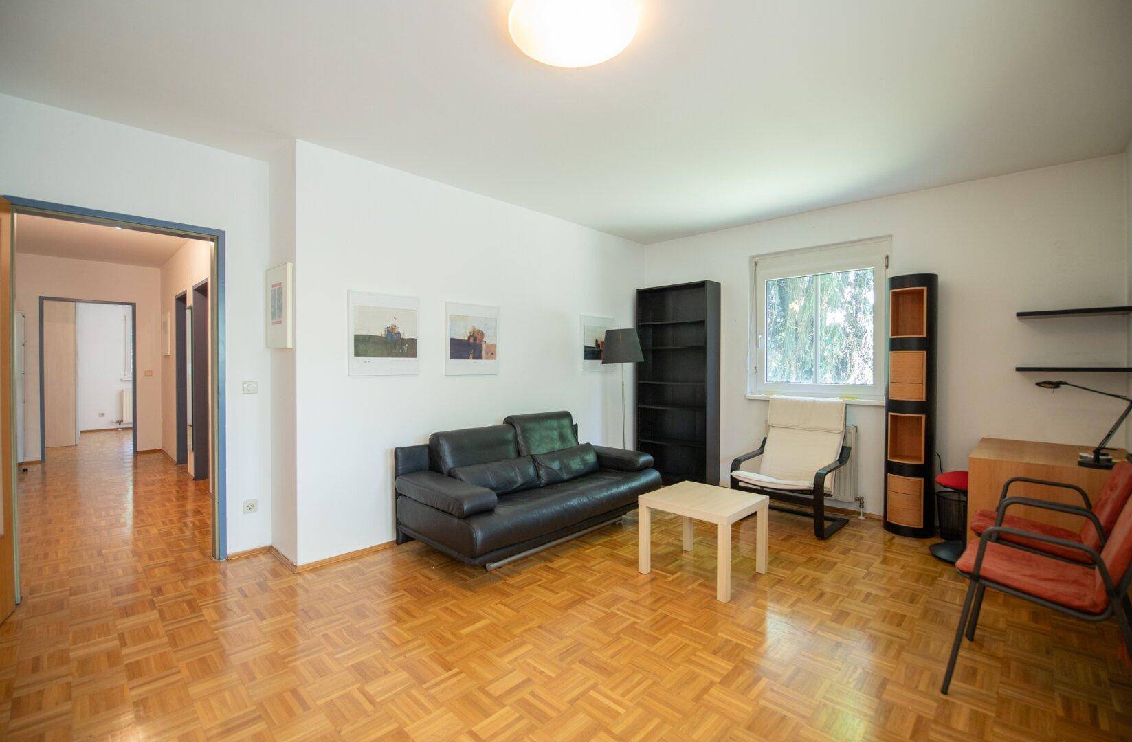Geräumiges Wohnzimmer mit Parkettboden, schwarzem Sofa und Blick in den angrenzenden Flur.