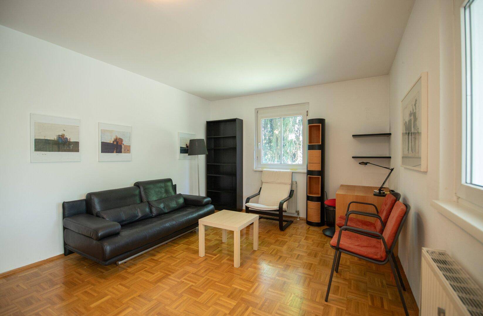 Helles Wohnzimmer mit Parkettboden, schwarzem Sofa und einem Fenster mit Blick ins Grüne.