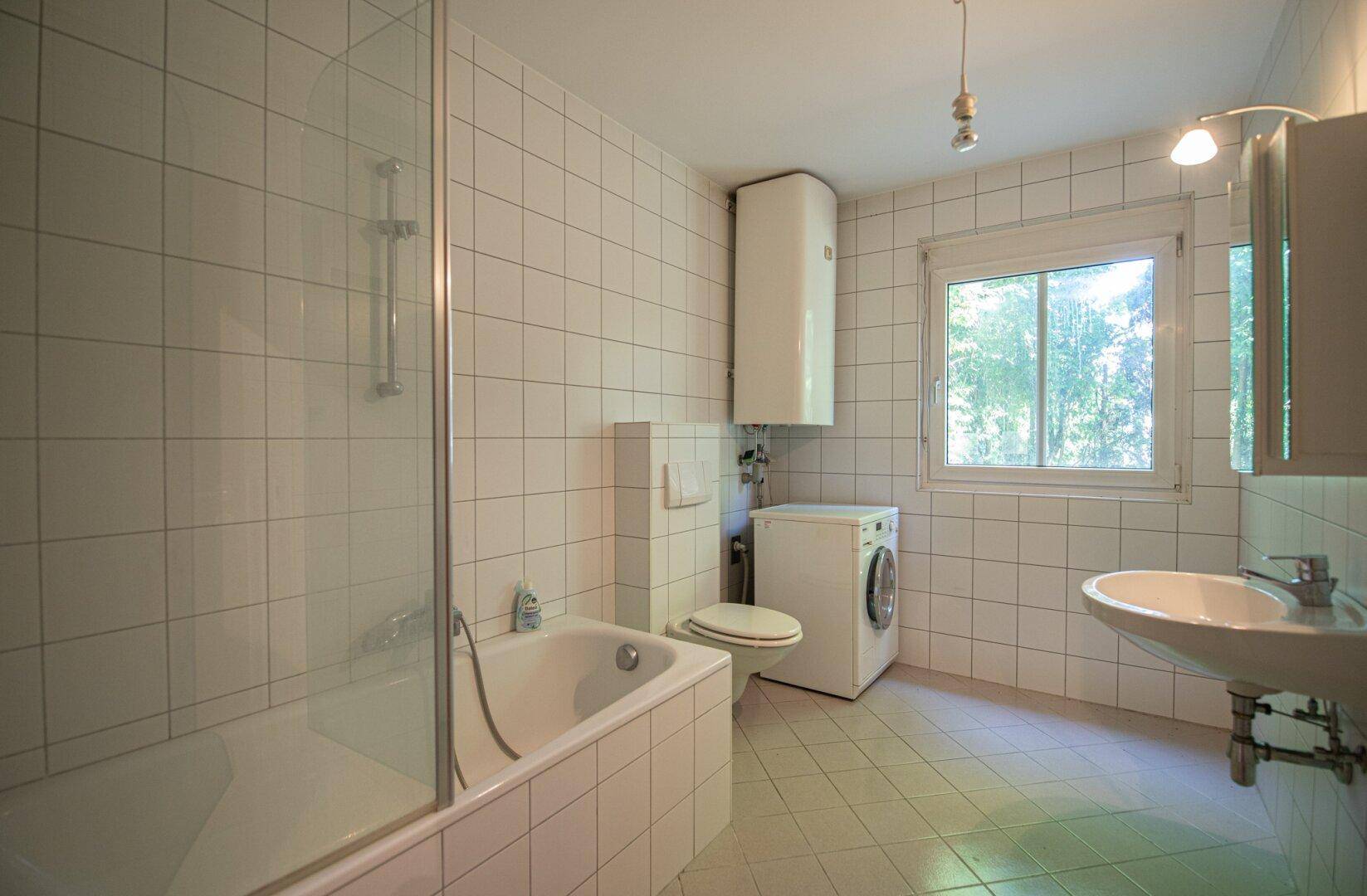 Gefliestes Badezimmer mit Badewanne, Toilette, Waschbecken und einem Fenster für Tageslicht.