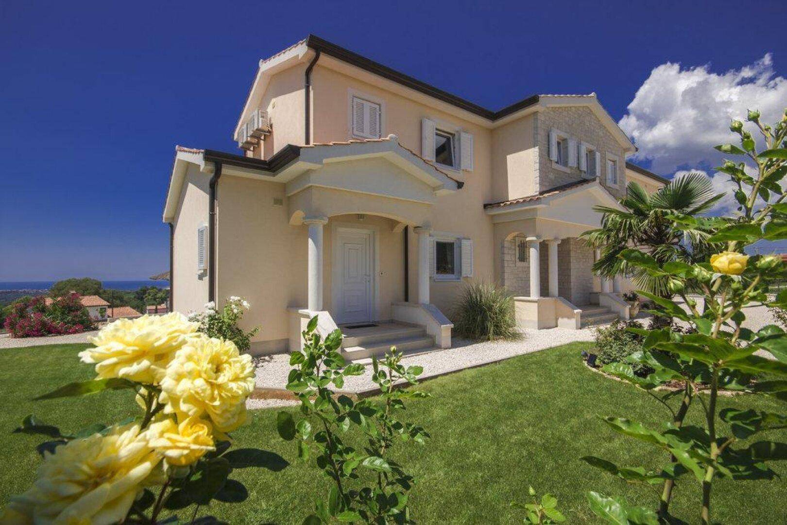 Eingangsbereich der Villa mit gepflegtem Rasen, gelben Rosen und einem weiten Blick auf das Meer.
