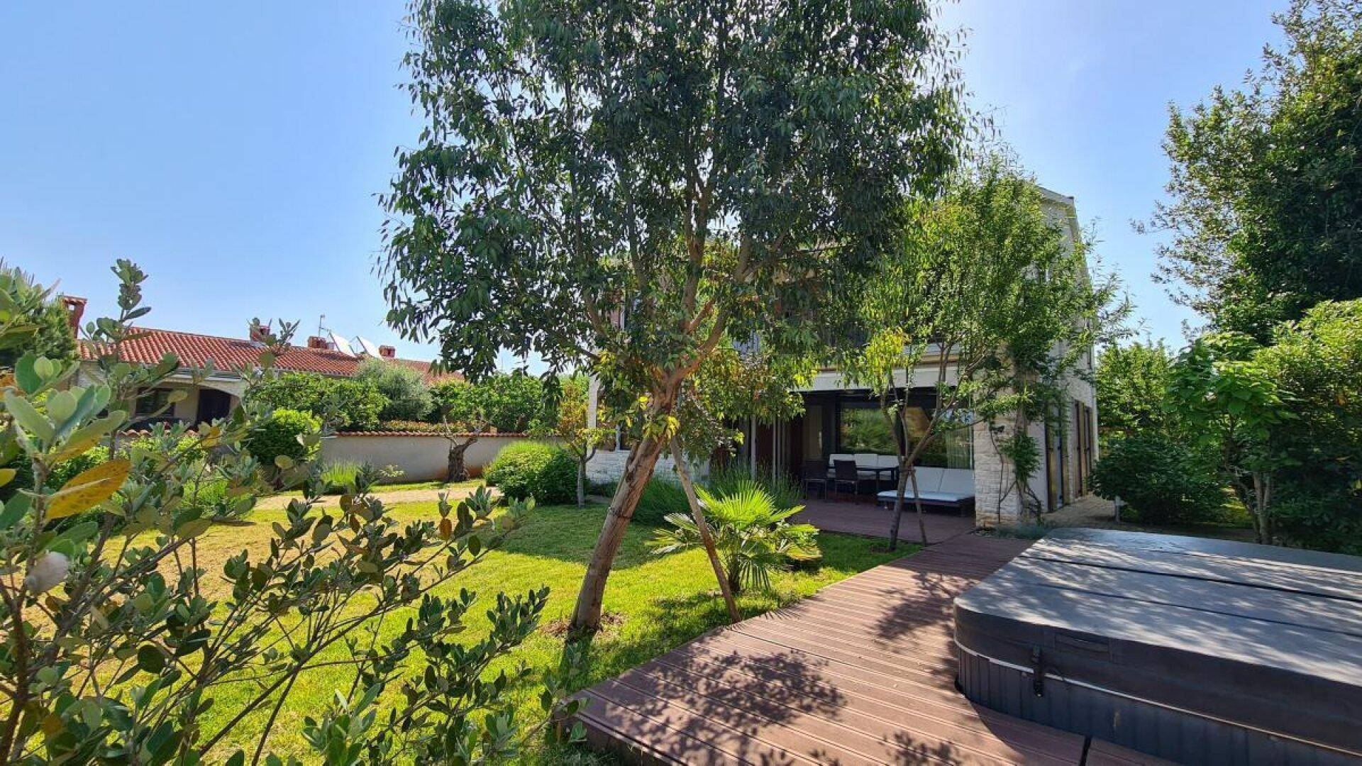 Großer Garten mit Holzdeck, Rasenfläche und einem Whirlpool, umgeben von Bäumen unter blauem Himmel.