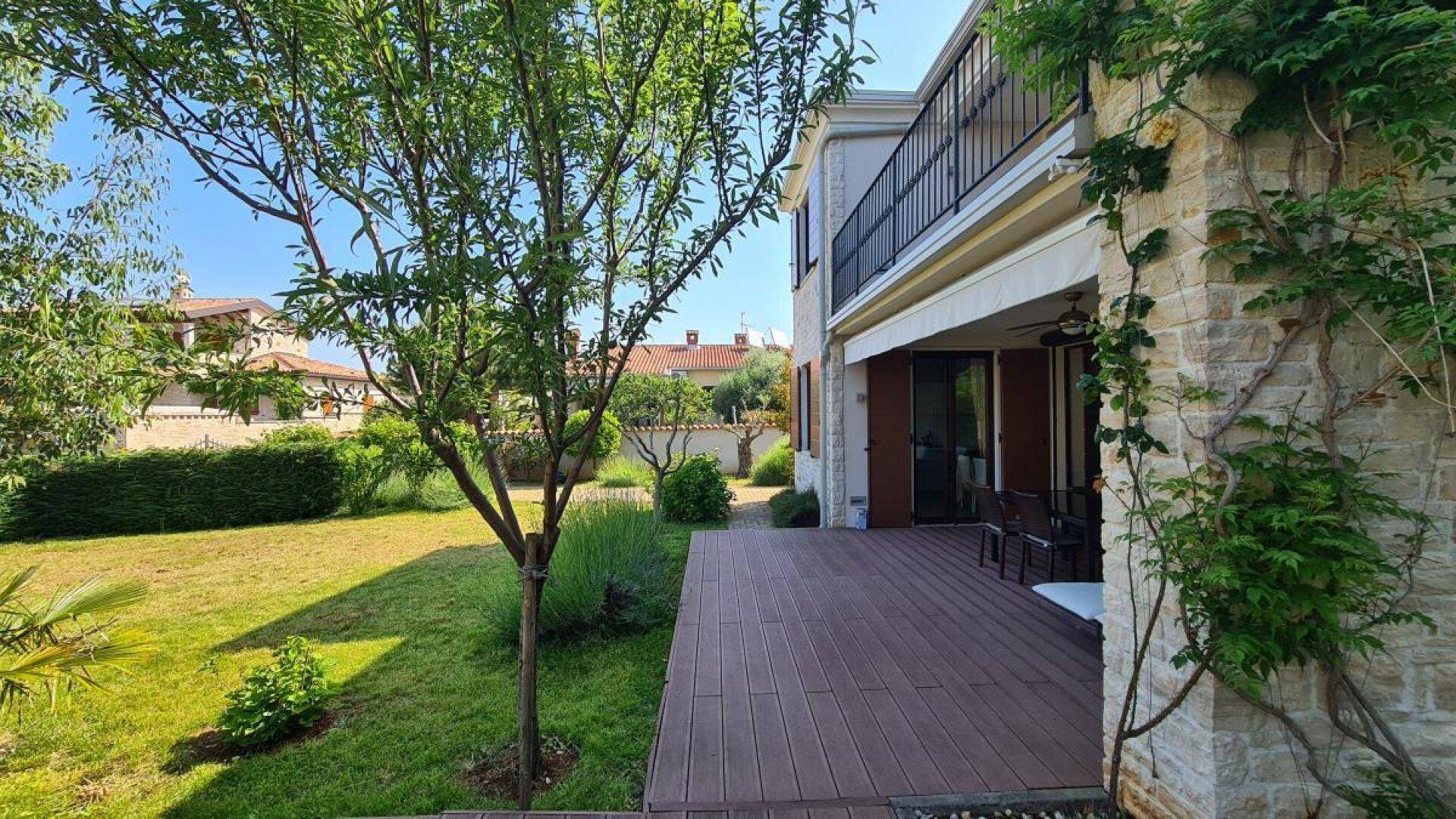 Seitlicher Blick auf das Haus mit einer Holzterrasse und einem weitläufigen Rasenbereich im Sonnenschein.