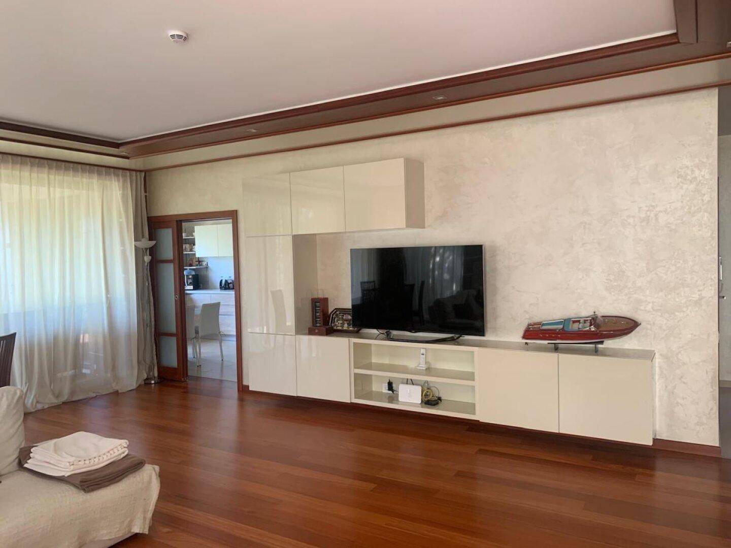 Modernes Wohnzimmer mit glänzender TV-Wand, Holzboden und Zugang zur Küche.