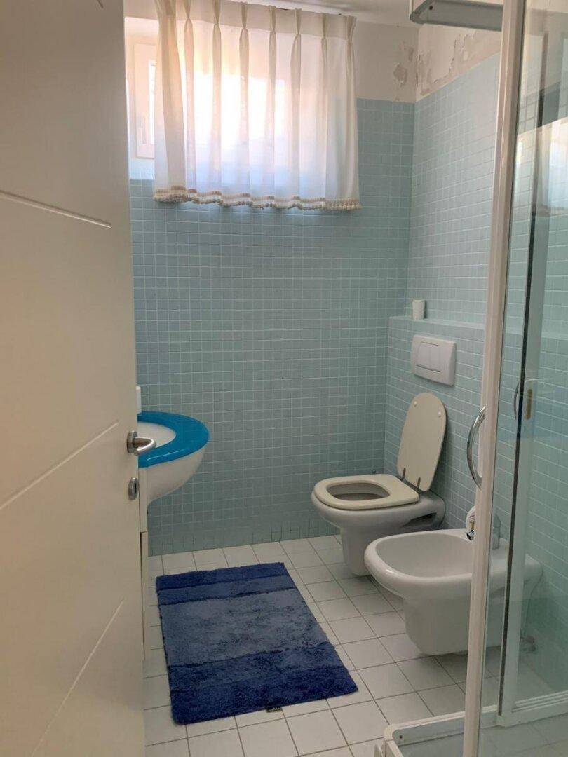 Badezimmer mit hellblauen Fliesen, Dusche, WC und Bidet, gut beleuchtet durch ein Fenster.