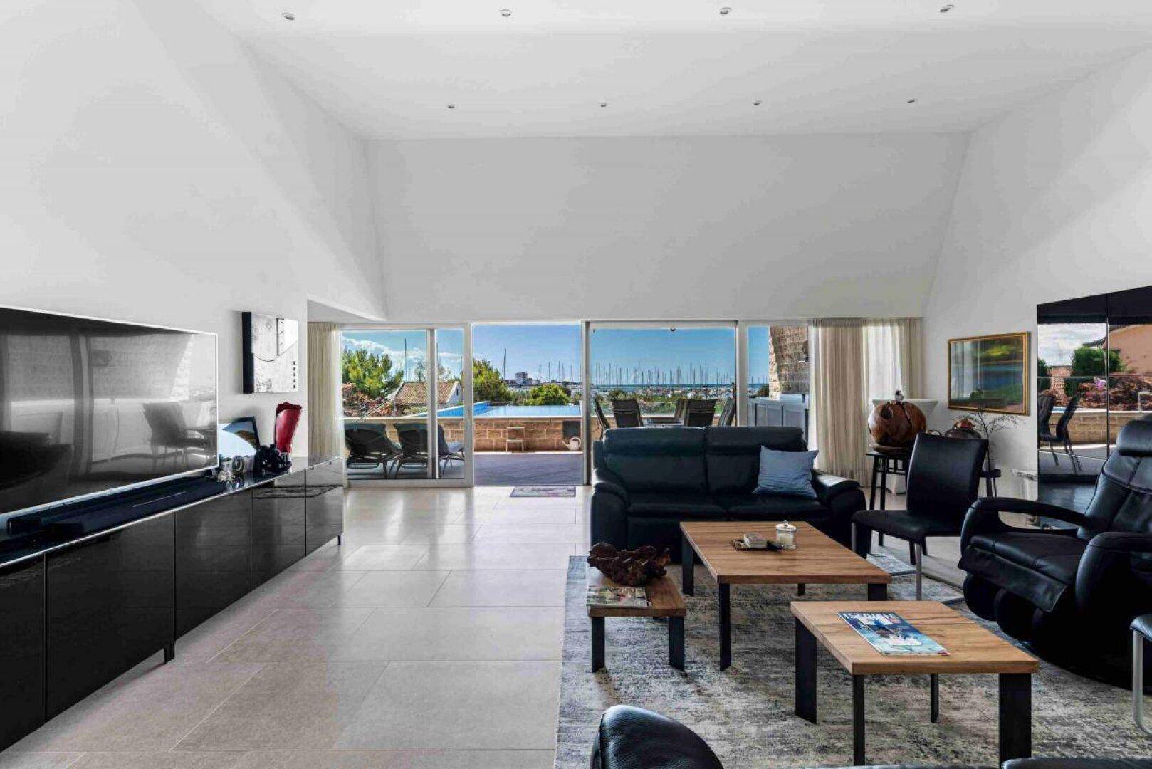 Helles Wohnzimmer mit großem Fernseher, schwarzen Möbeln und weitem Blick auf den Pool und das Meer.