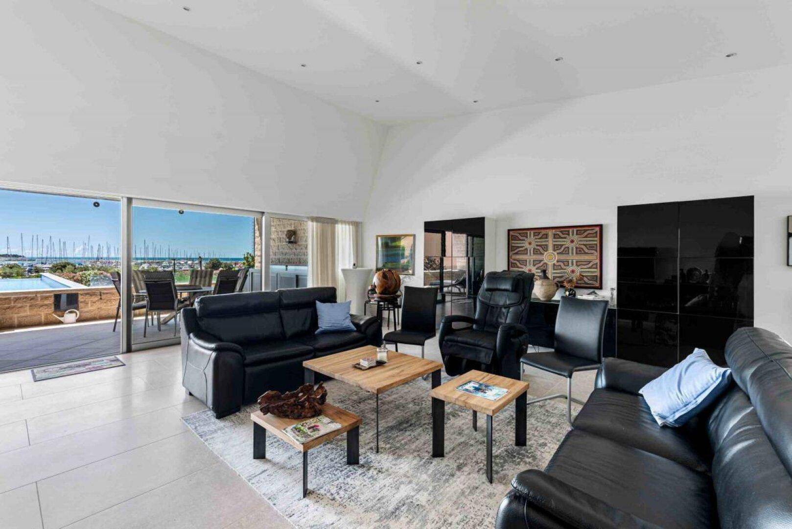 Geräumiges Wohnzimmer mit schwarzen Ledersofas, Holztischen und Zugang zur Terrasse mit Meerblick.