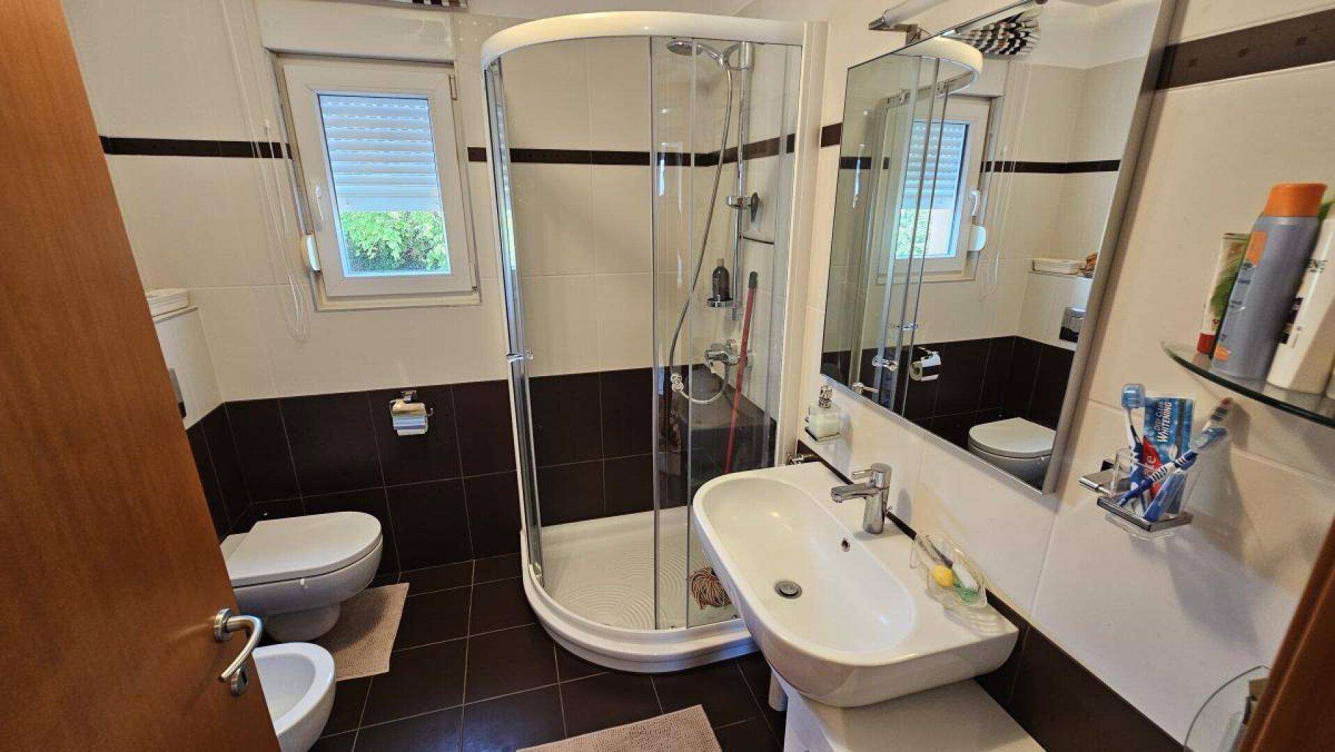 Badezimmer mit Duschkabine, Waschbecken, WC, Bidet und Fenster, in zweifarbigen Fliesen.