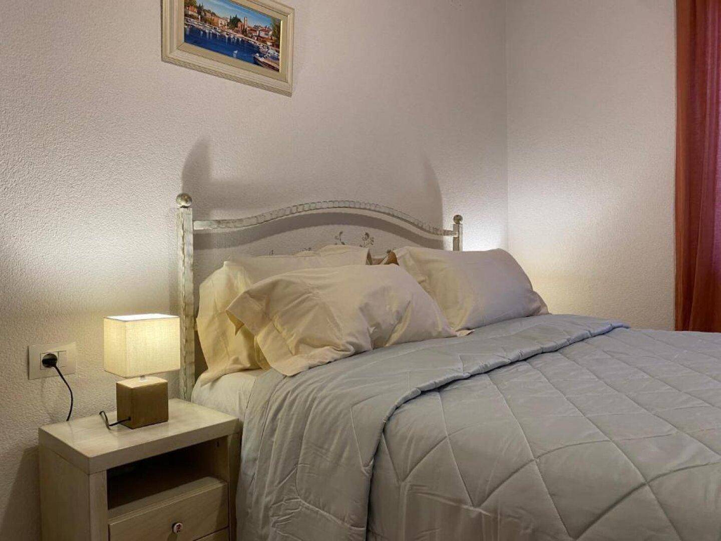 Helles Schlafzimmer mit einem schmiedeeisernen Bett, Nachttisch und einer kleinen Lampe.