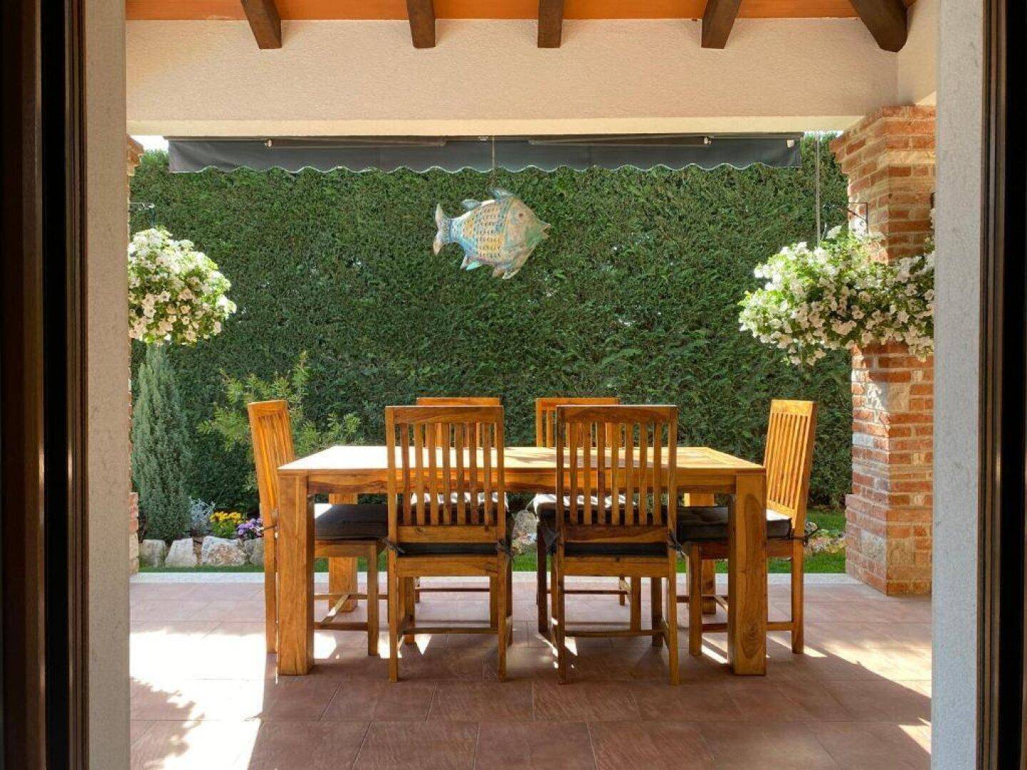 Überdachte Terrasse mit Holztisch und Stühlen, umgeben von einer grünen Hecke.