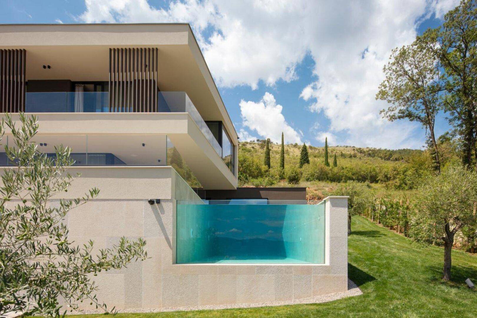 Seitliche Ansicht der modernen Villa mit einzigartigem Glas-Infinity-Pool und grüner Umgebung.