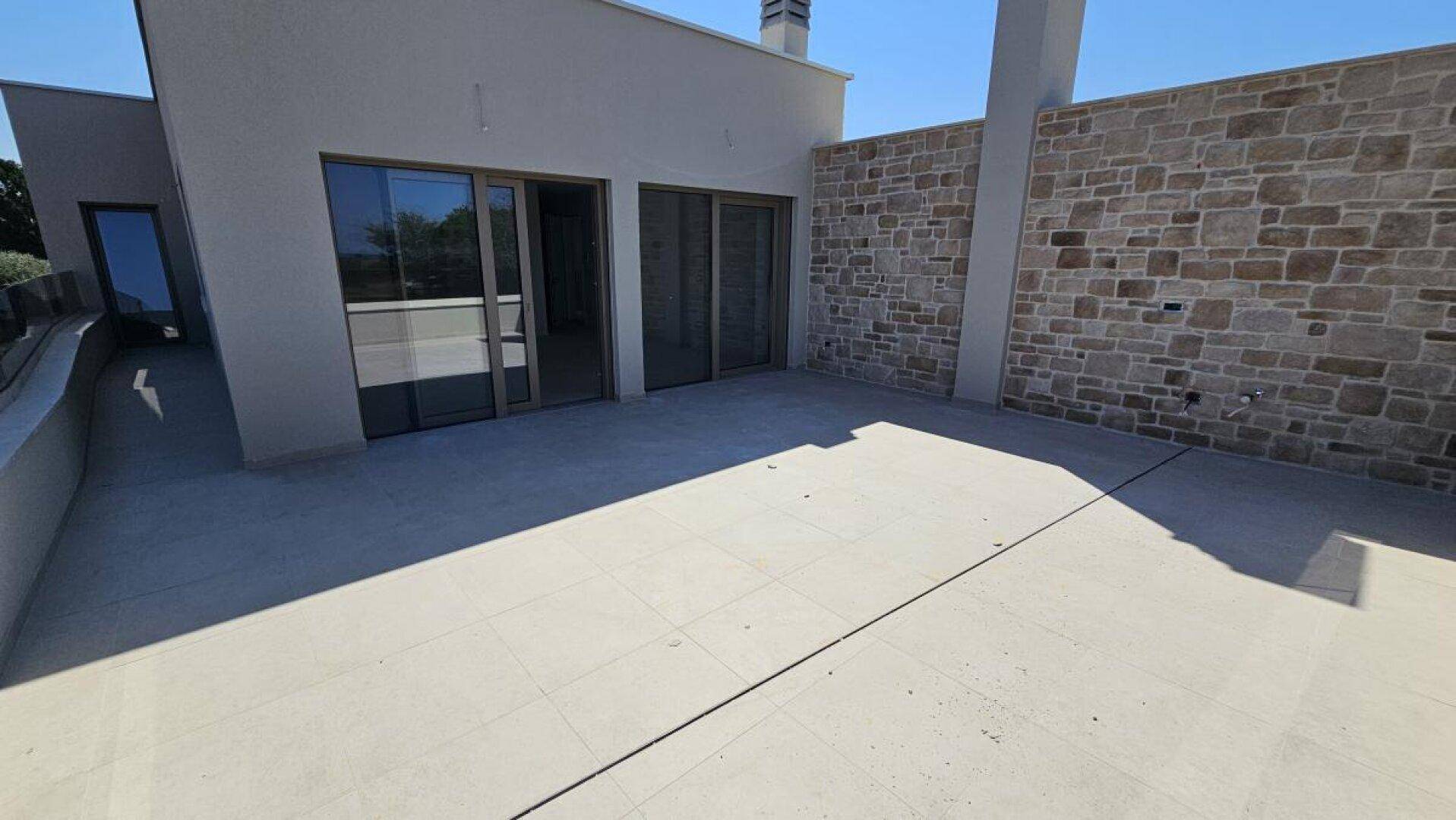 Große geflieste Terrasse mit Steinwand und Zugang zu den Innenräumen des modernen Gebäudes.