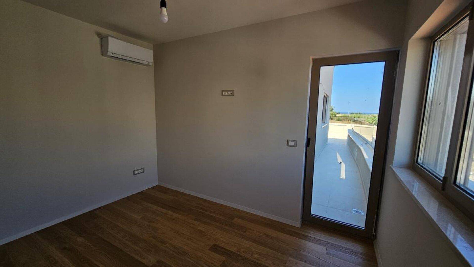 Heller Raum mit Holzdielenboden, Klimaanlage und Zugang zu einem Außenbereich mit Meerblick.