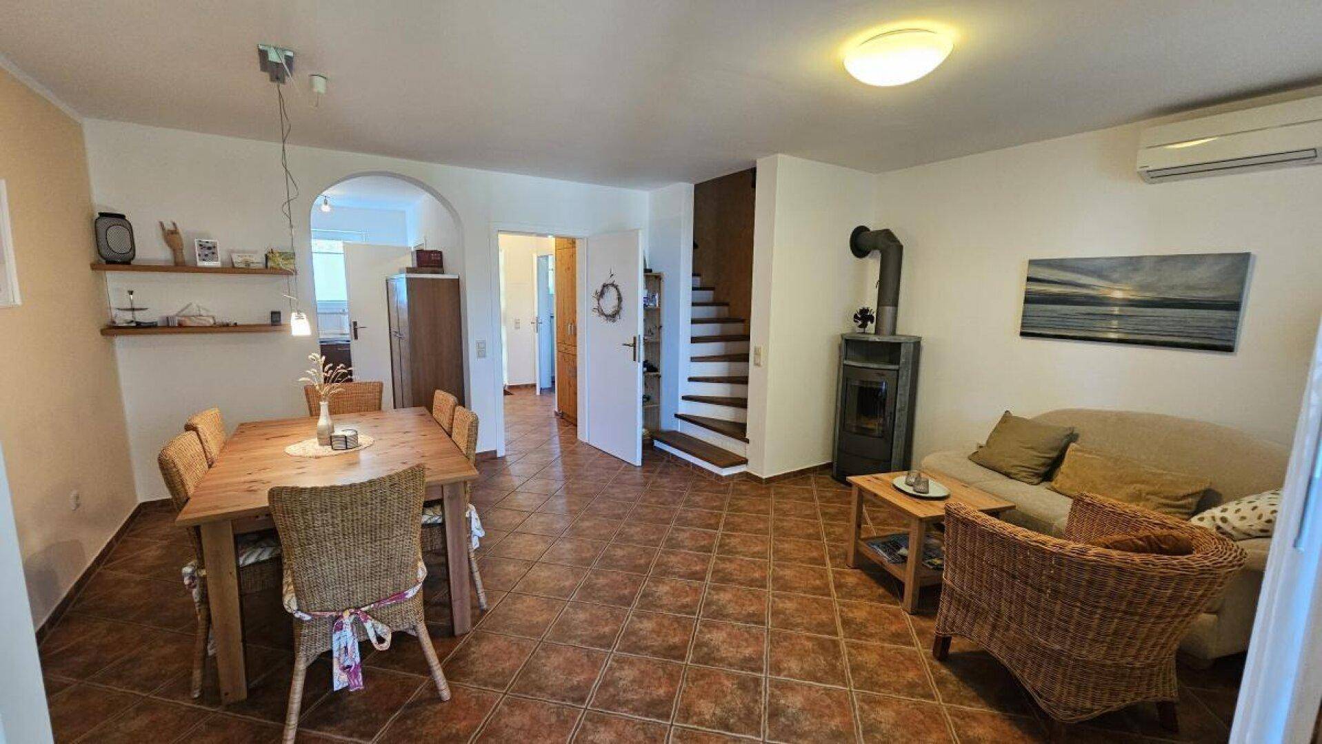 Gemütlicher Wohn-Essbereich mit Kaminofen, Fliesenboden und Zugang zu einer Treppe.