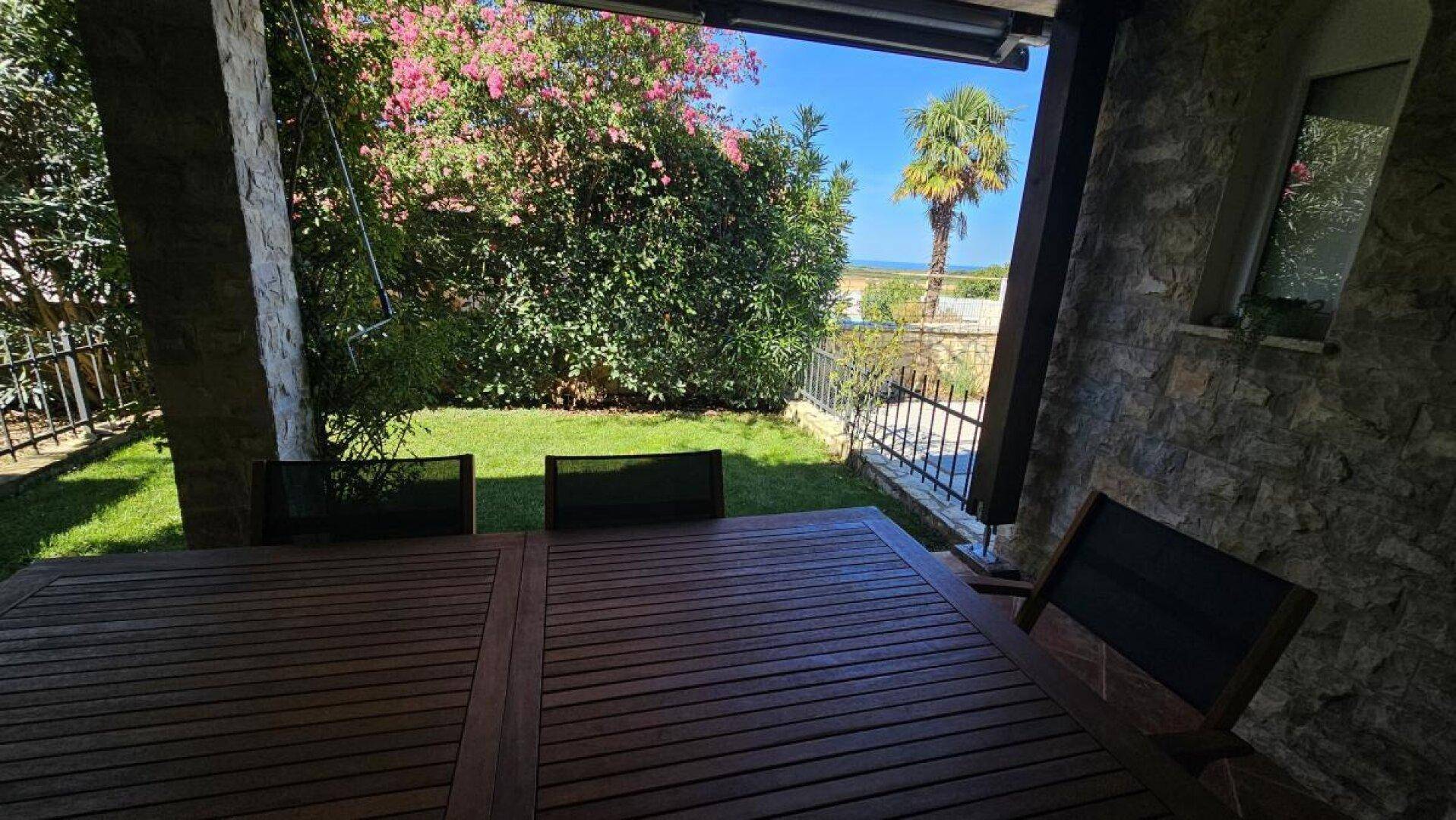 Überdachte Terrasse mit Holztisch und Gartenblick, ideal für Mahlzeiten im Freien mit Meer im Hintergrund.