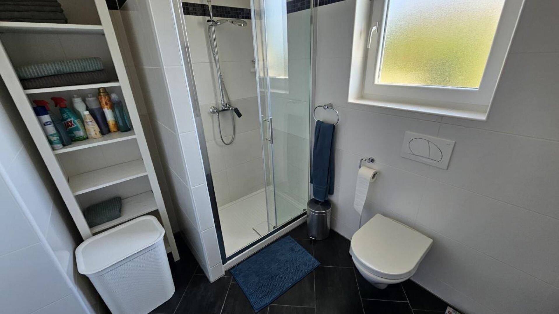 Helles Badezimmer mit Duschkabine, WC, Fenster und praktischem Regal für Badutensilien.