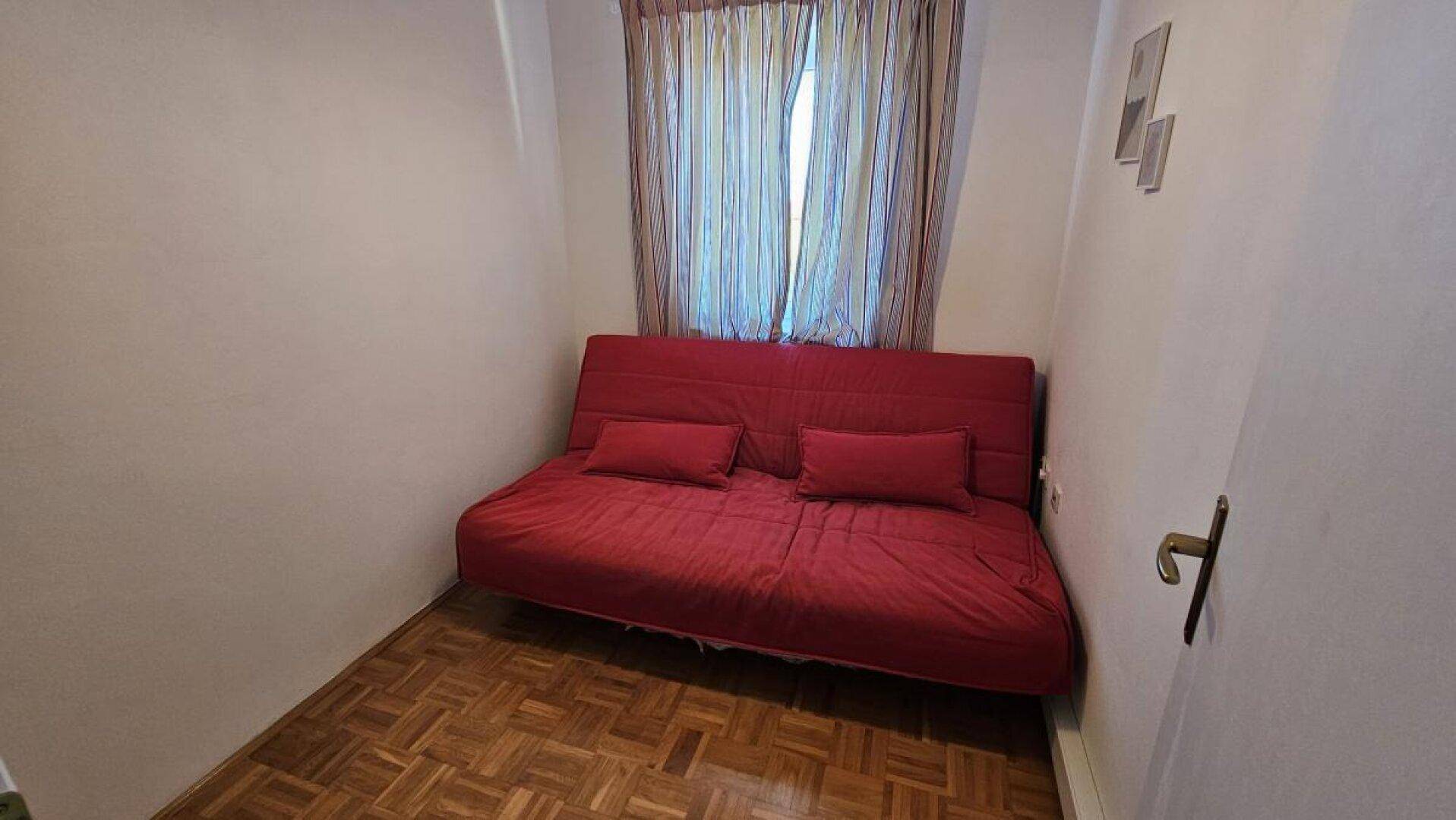 Kleines Zimmer mit rotem Schlafsofa und Parkettboden, ideal als Gästezimmer oder Arbeitsbereich.
