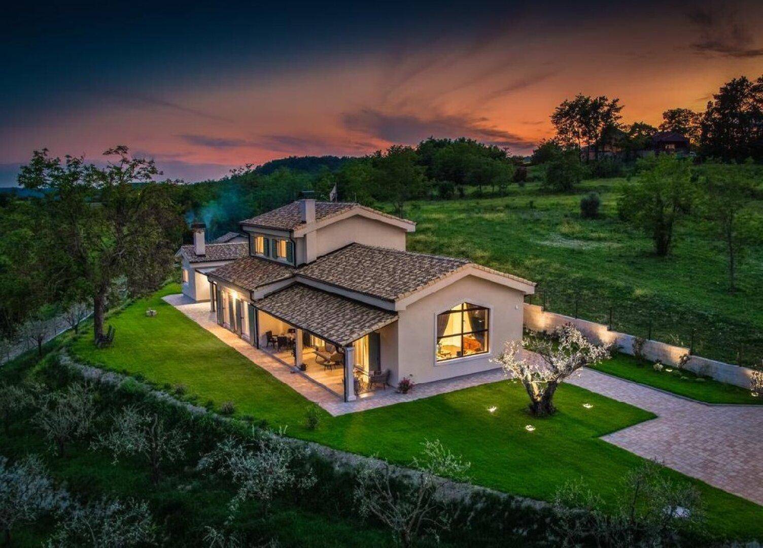 Panoramablick auf eine elegante Villa bei Sonnenuntergang, umgeben von grüner Landschaft und einem Pool.