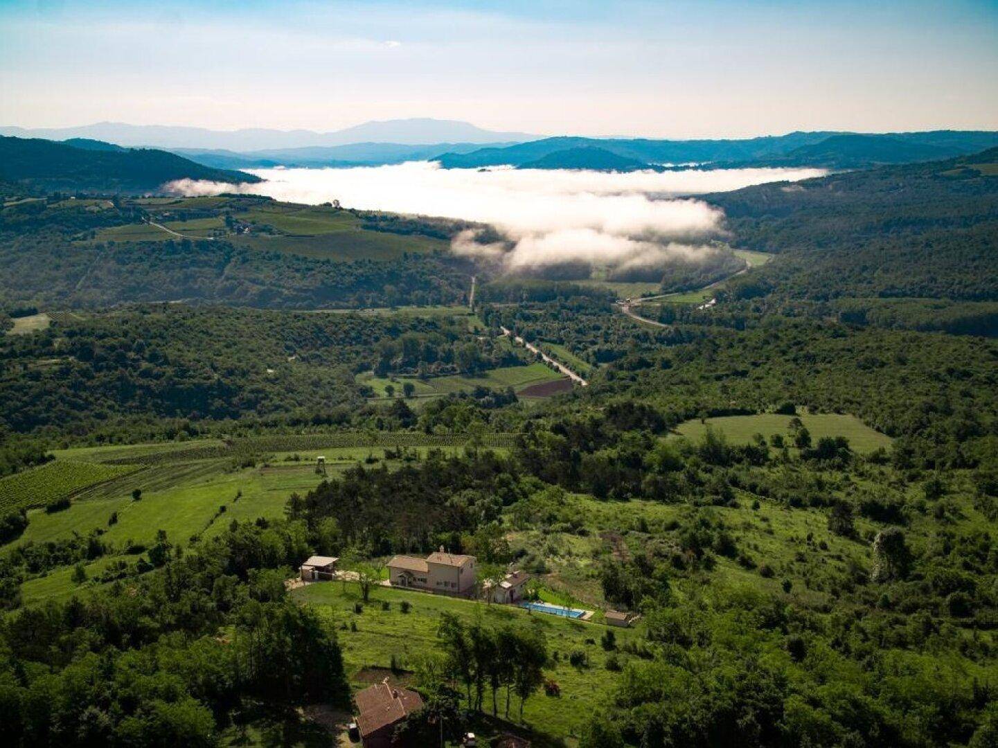 Weitläufige Luftaufnahme der Villa inmitten einer hügeligen, grünen Landschaft mit Nebel in der Ferne.