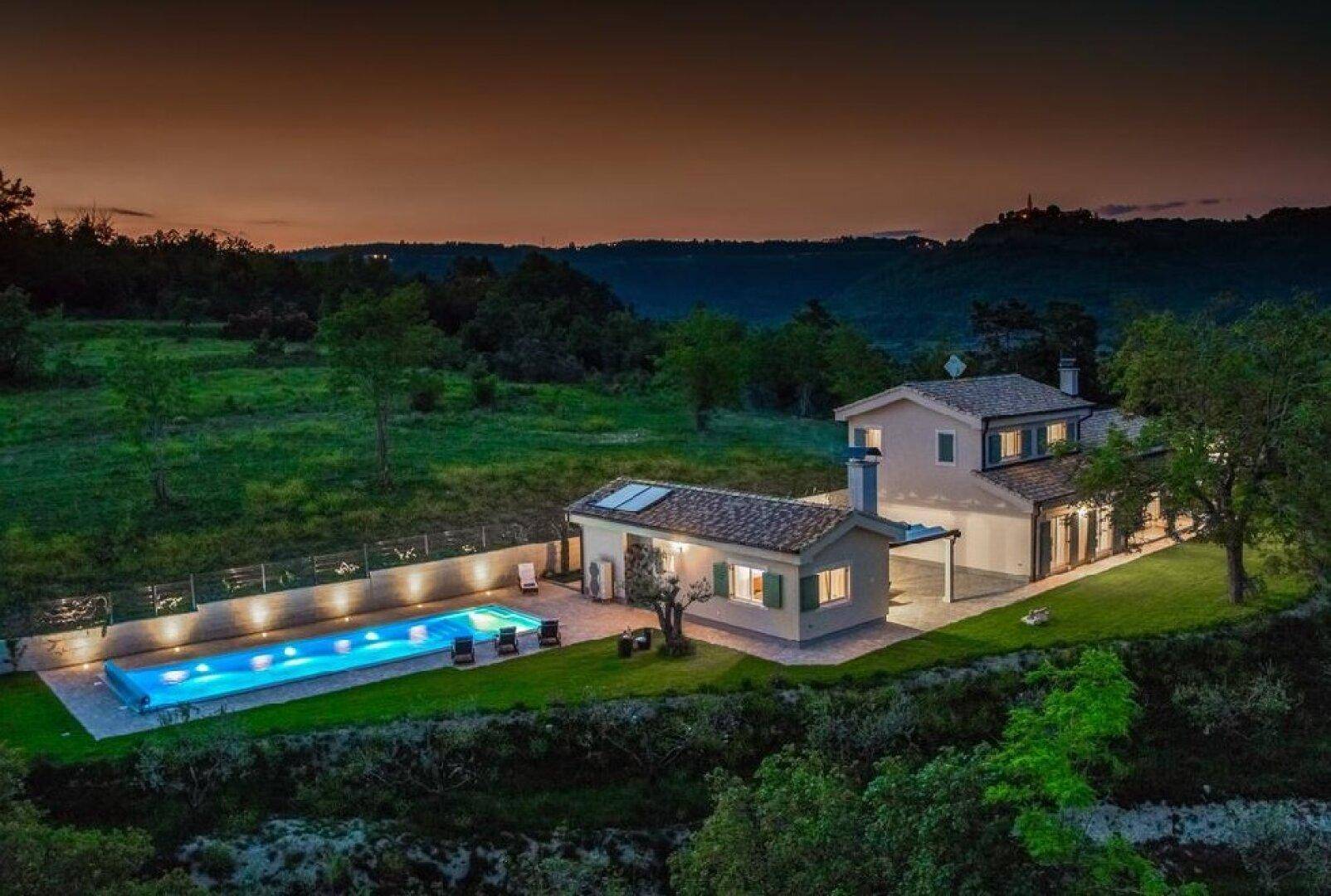 Abendliche Luftaufnahme einer eleganten Villa mit beleuchtetem Pool und weitläufigem Grundstück in der Dämmerung.