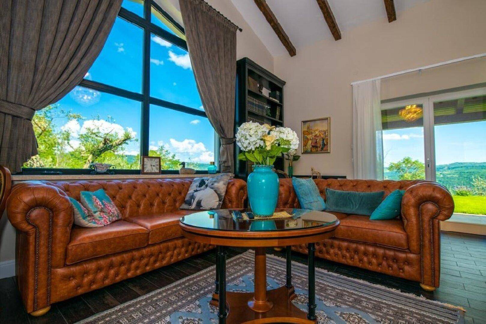Gemütliches Wohnzimmer mit großen Fenstern, Ledersofas, Holzbalken und Blick ins Grüne.