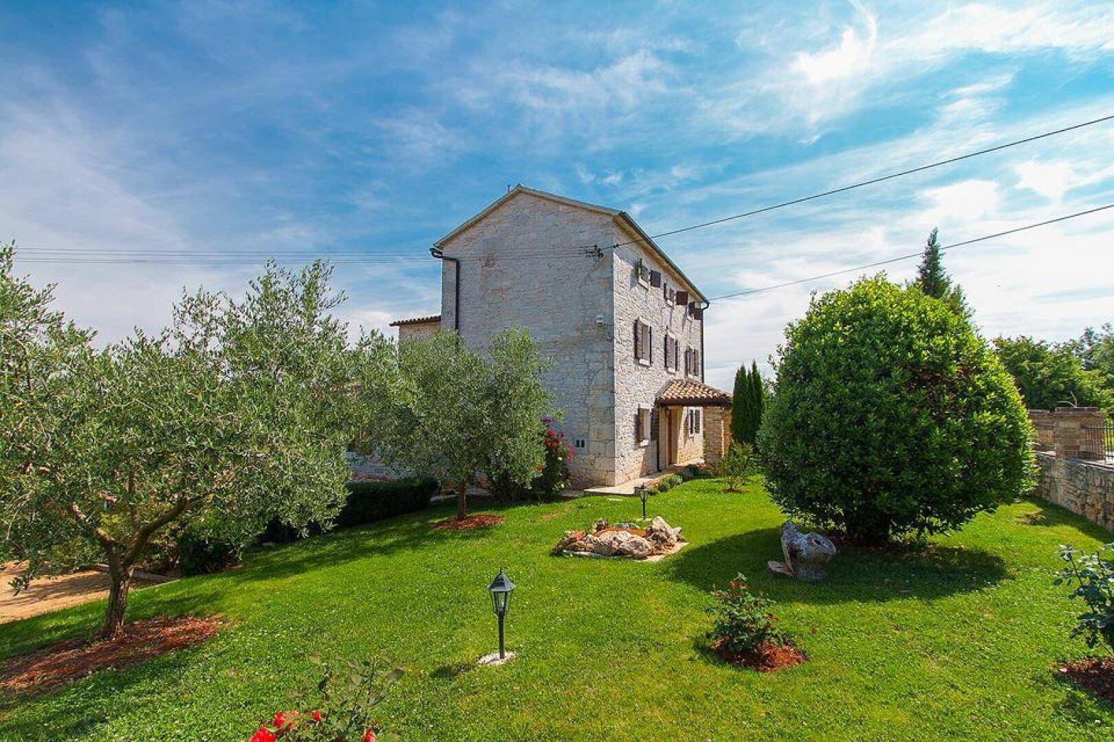 Seitlicher Blick auf das traditionelle Steinhaus mit gepflegtem Garten, Olivenbäumen und grüner Rasenfläche.