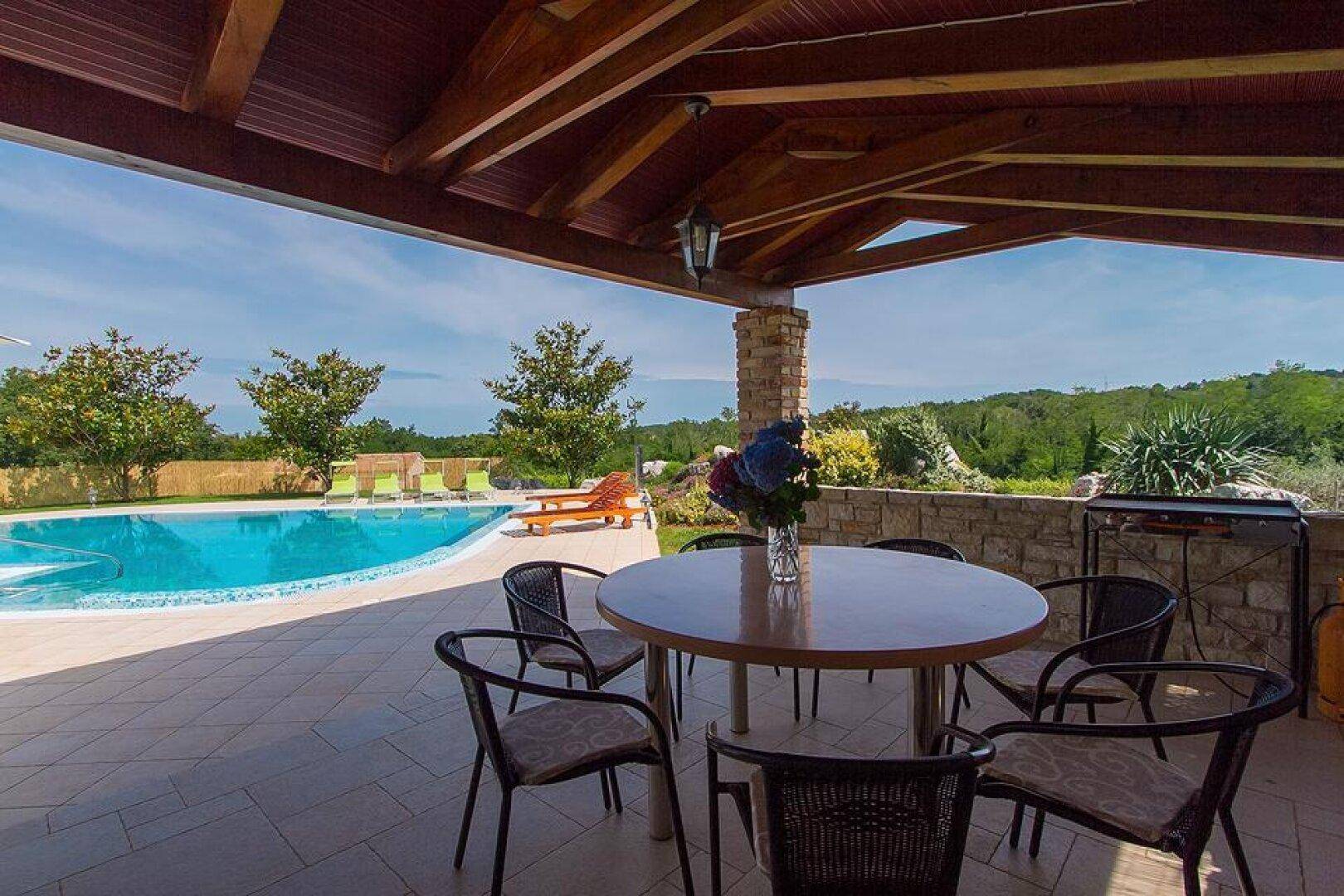 Gemütlicher überdachter Essbereich im Freien mit Grill und Blick auf den Pool und die grüne Landschaft.