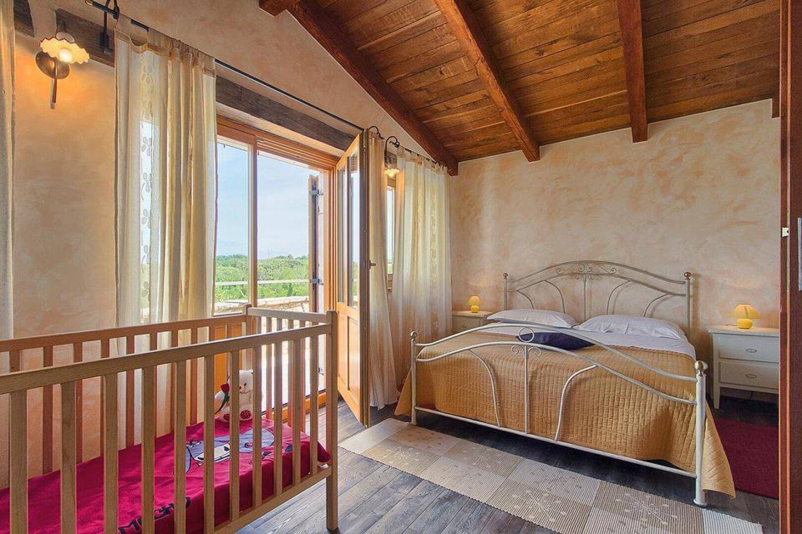 Helles Schlafzimmer mit Holzbalkendecke, Doppelbett und Babybett, mit Zugang zu einem französischen Balkon.