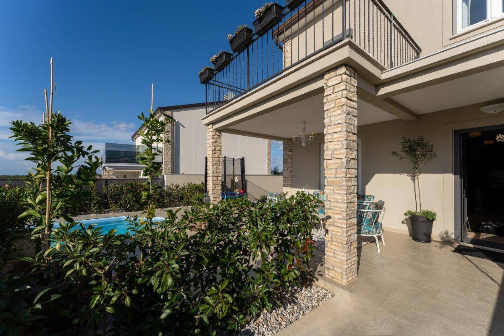 Ansicht des Hauses mit überdachter Terrasse, umgeben von grünen Pflanzen und einem Pool.