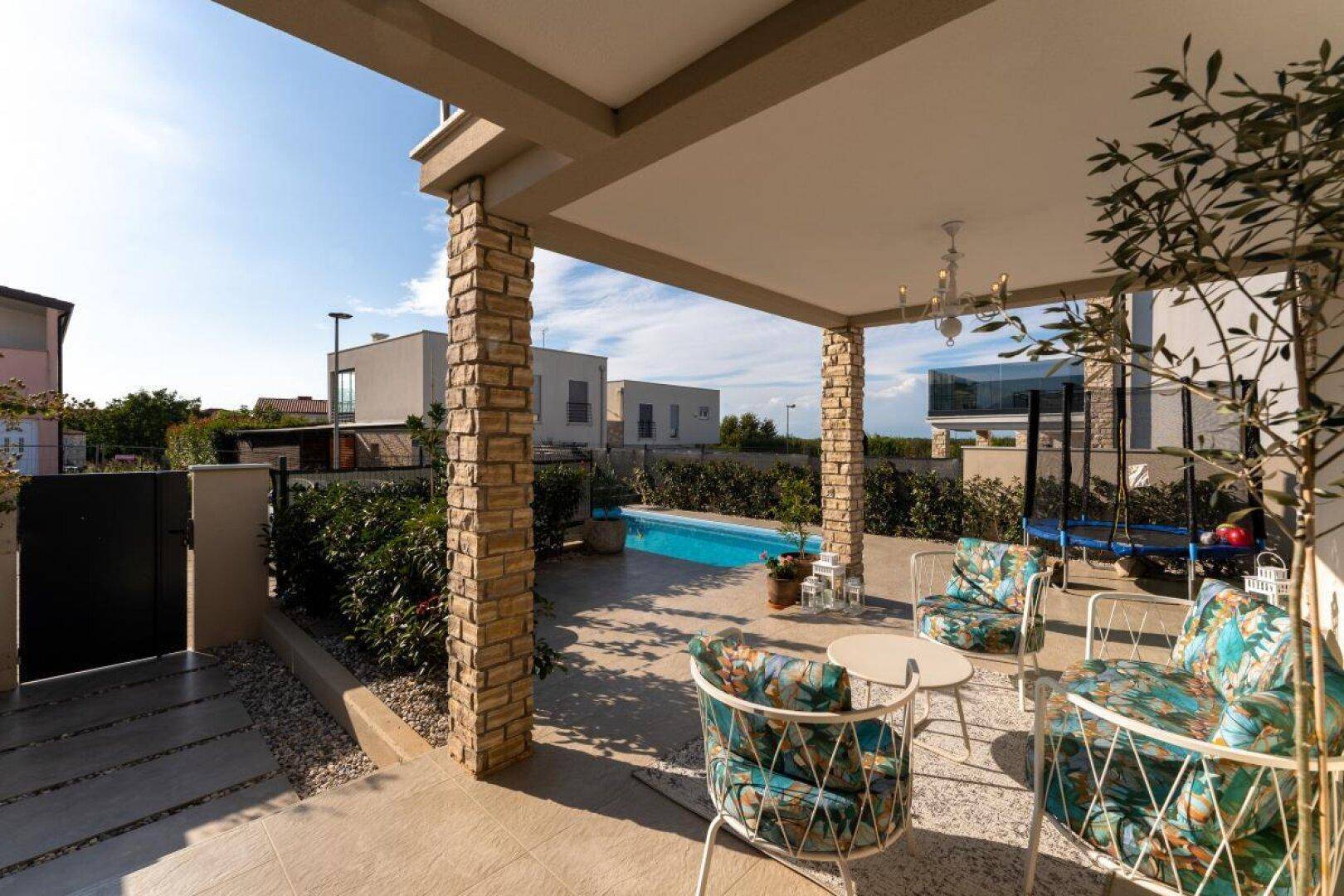 Überdachte Terrasse mit Steinpfeilern, Sitzgelegenheiten und Blick auf den Garten mit Pool.
