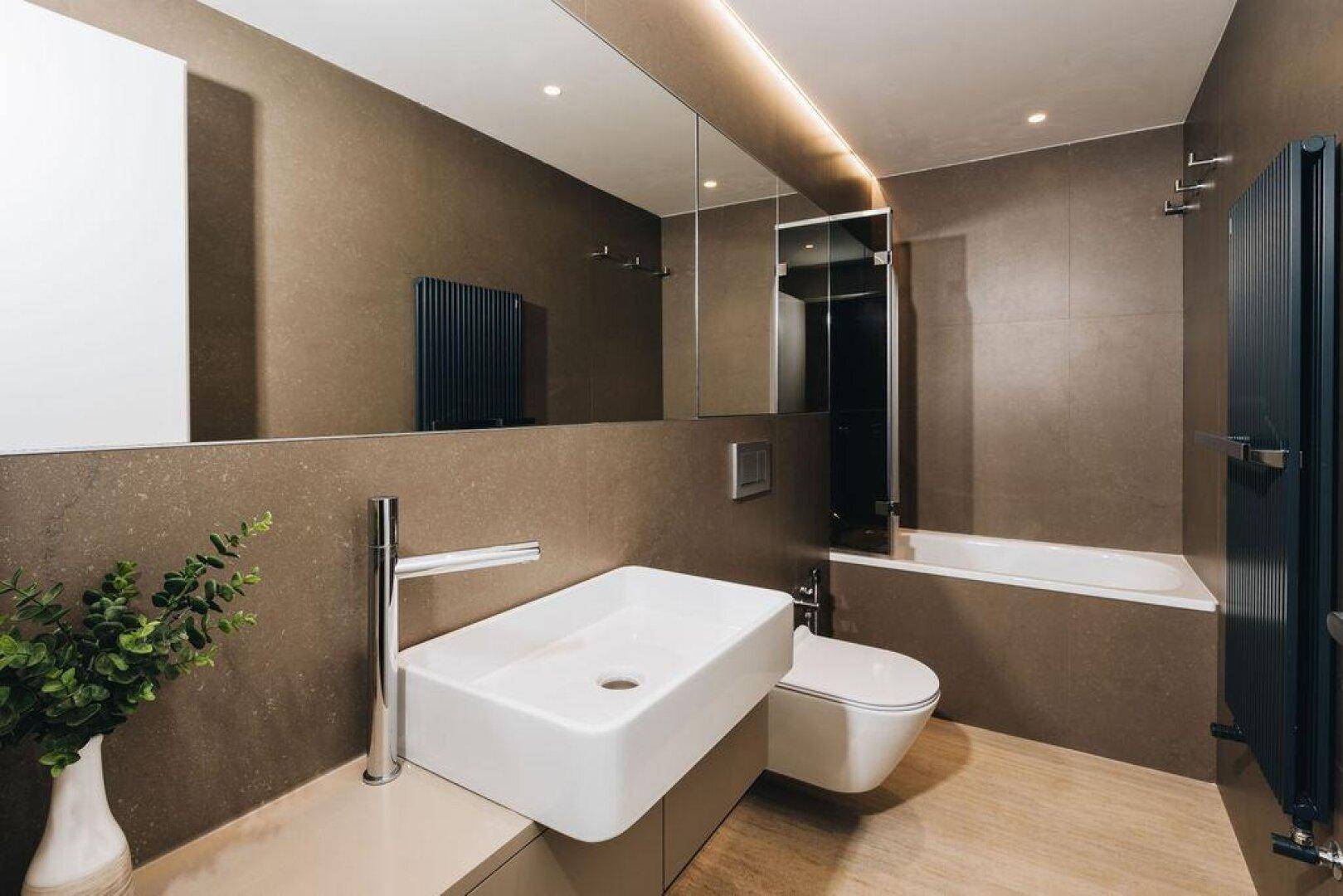Elegantes Badezimmer mit breitem Waschtisch, integriertem Spiegel und dunklen Fliesen.