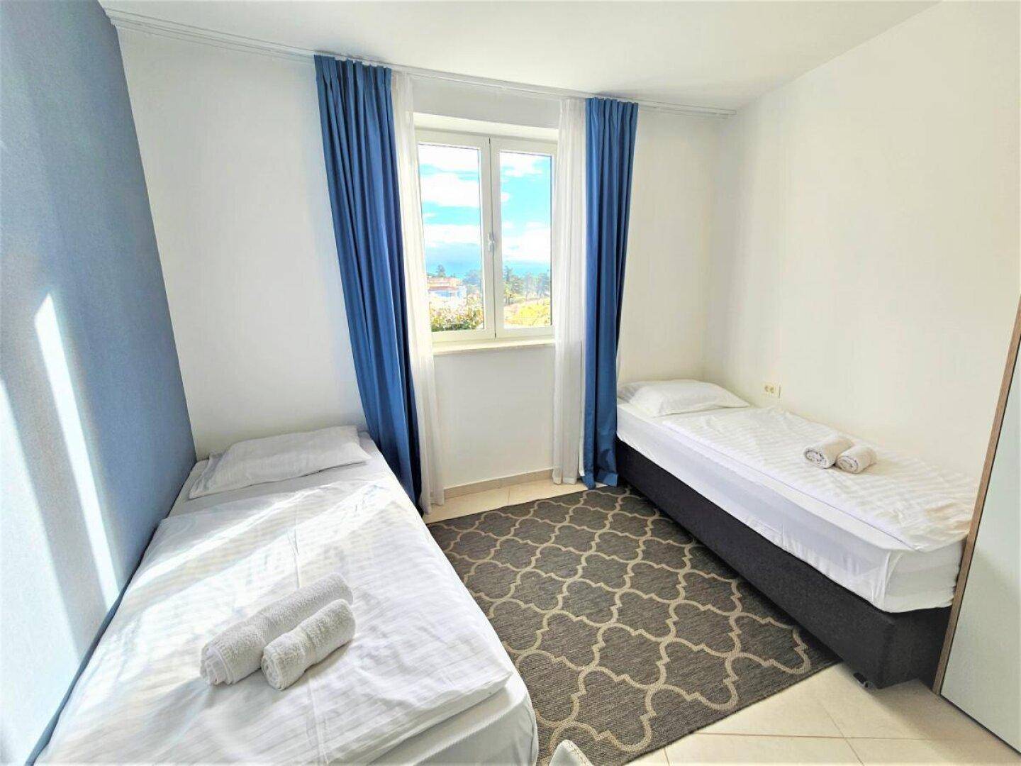 Schlafzimmer mit zwei Einzelbetten, blauen Vorhängen und einem Fenster, das Tageslicht hereinlässt.