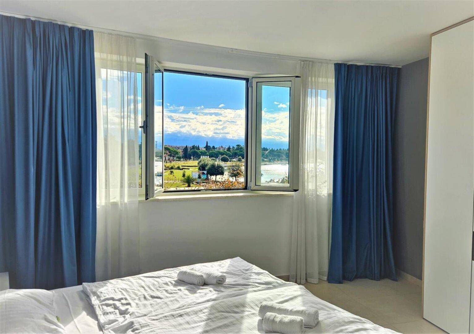 Helles Schlafzimmer mit Doppelbett, blauen Vorhängen und einem Fenster mit Blick auf die Bucht.
