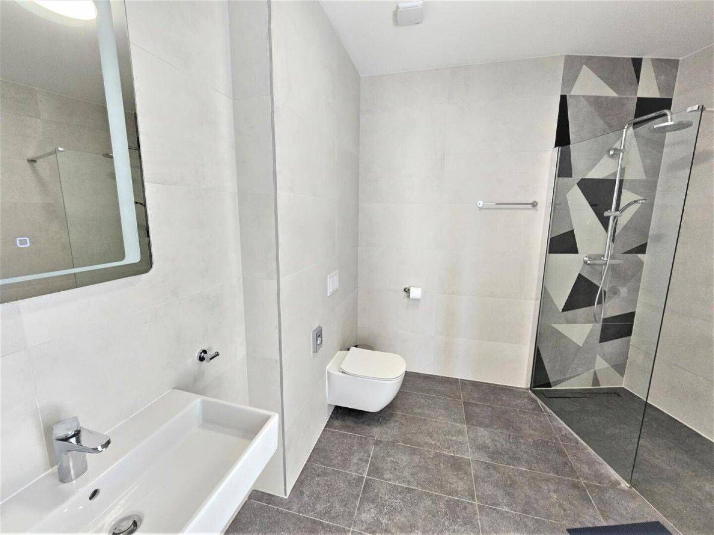 Modernes Badezimmer mit hellgrauen Fliesen, wandhängendem WC und einer begehbaren Dusche mit geometrischem Muster.