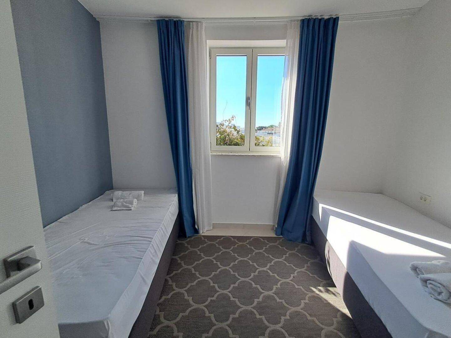 Schlafzimmer mit zwei Einzelbetten, blauen Vorhängen und einem Fenster, das den Raum erhellt.