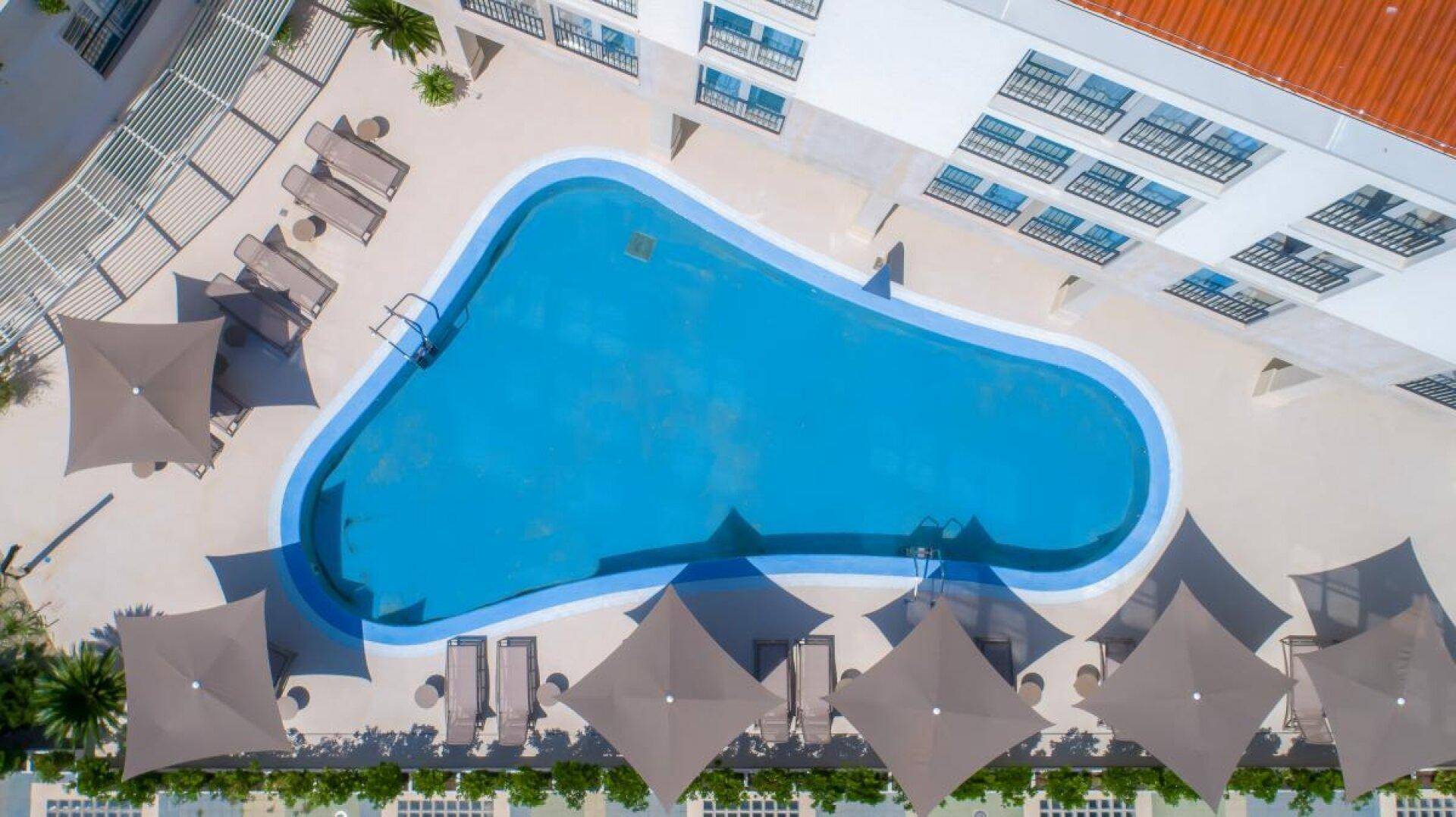 Luftaufnahme eines großen Pools mit Liegestühlen und Sonnenschirmen, umgeben von einem Gebäude.