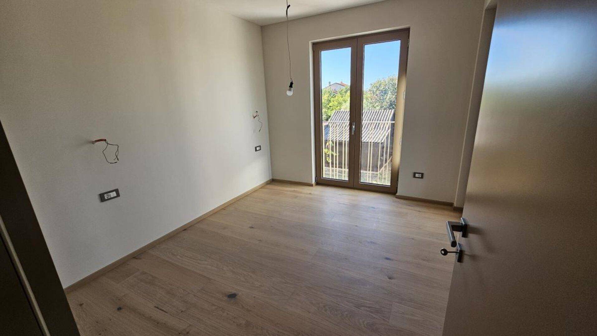 Helles Schlafzimmer mit Holzboden und Fenstertür, die Zugang zu einem kleinen Balkon bietet.