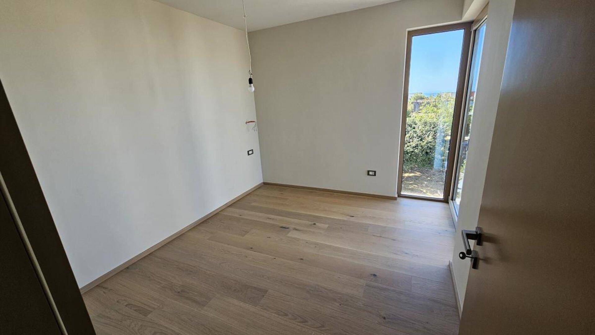 Helles Schlafzimmer mit Holzboden und großem Fenster, das einen ruhigen Blick ins Grüne bietet.