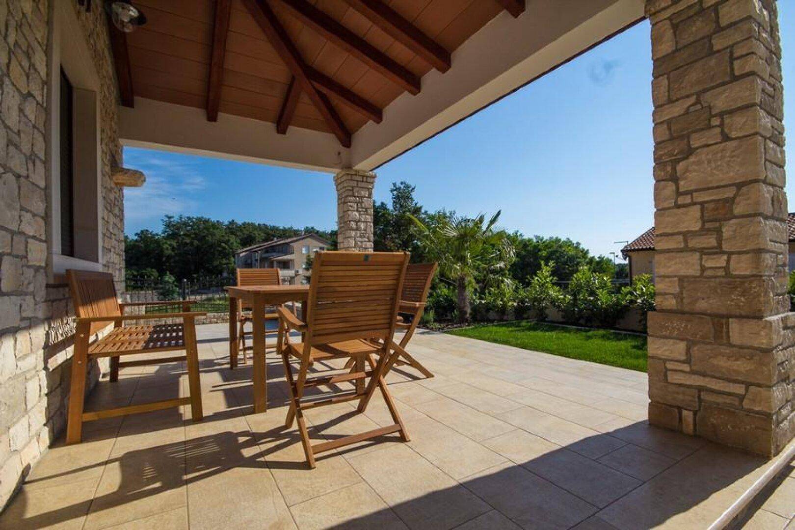 Überdachte Terrasse mit Holztisch und Stühlen, umgeben von Steinmauern und grüner Natur.