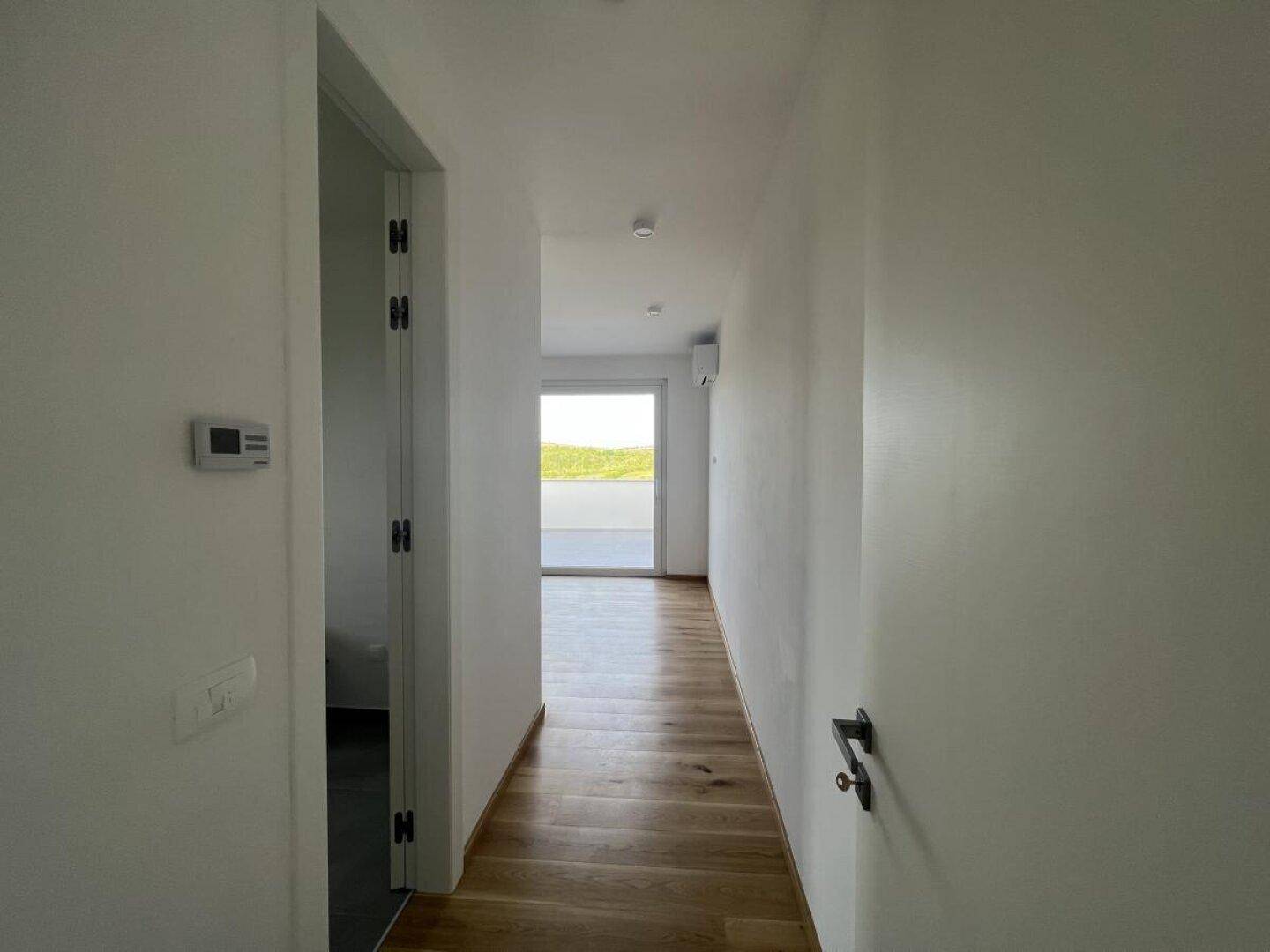 Langer, heller Flur mit Holzboden, der zu einem Zimmer mit Balkonzugang führt.