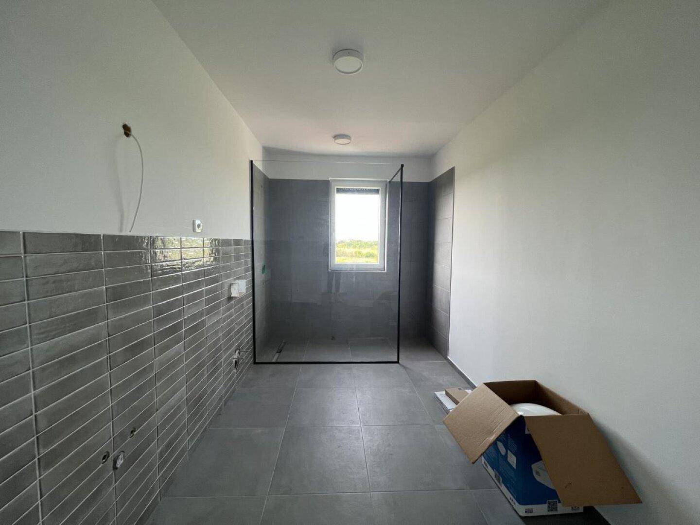 Geräumiges Badezimmer im Bau mit grauen Fliesen, Fenster und begehbarer Dusche.